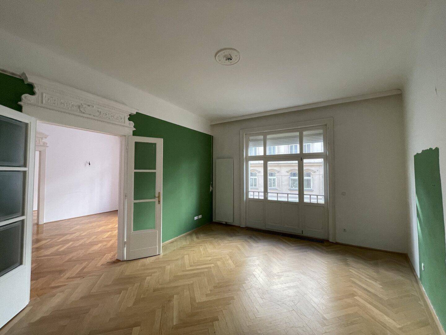 Wohnzimmer mit grüner Akzentwand, Fischgrätparkett und großen Fenstern.