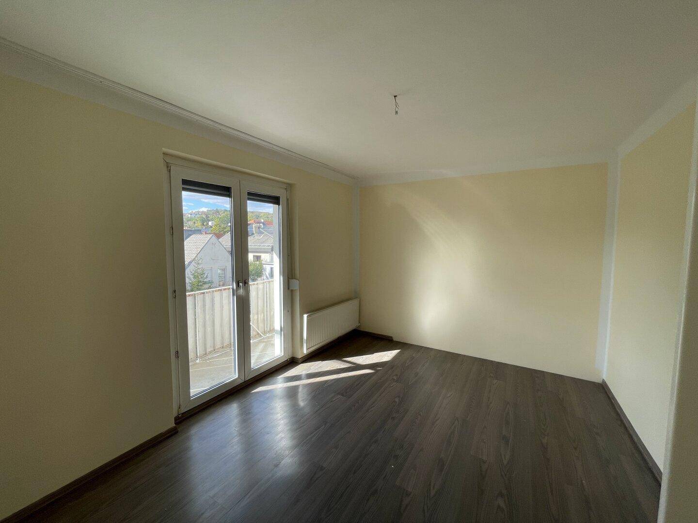 Helles, leeres Zimmer mit dunklem Laminatboden und Zugang zu einem Balkon, gut beleuchtet.