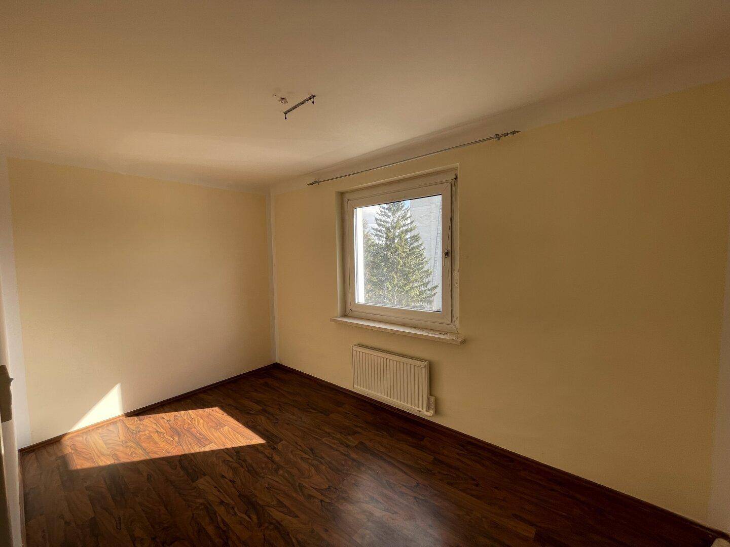 Helles, leeres Zimmer mit dunklem Laminatboden und einem Fenster mit Blick auf Bäume.