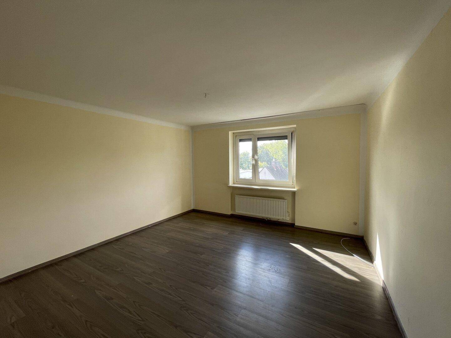 Leeres Zimmer mit Laminatboden und einem Fenster, das natürliches Licht in den Raum bringt.