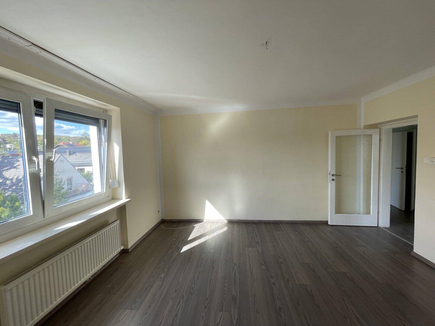 Heller, leerer Raum mit Laminatboden und großem Fenster, das viel natürliches Licht spendet.