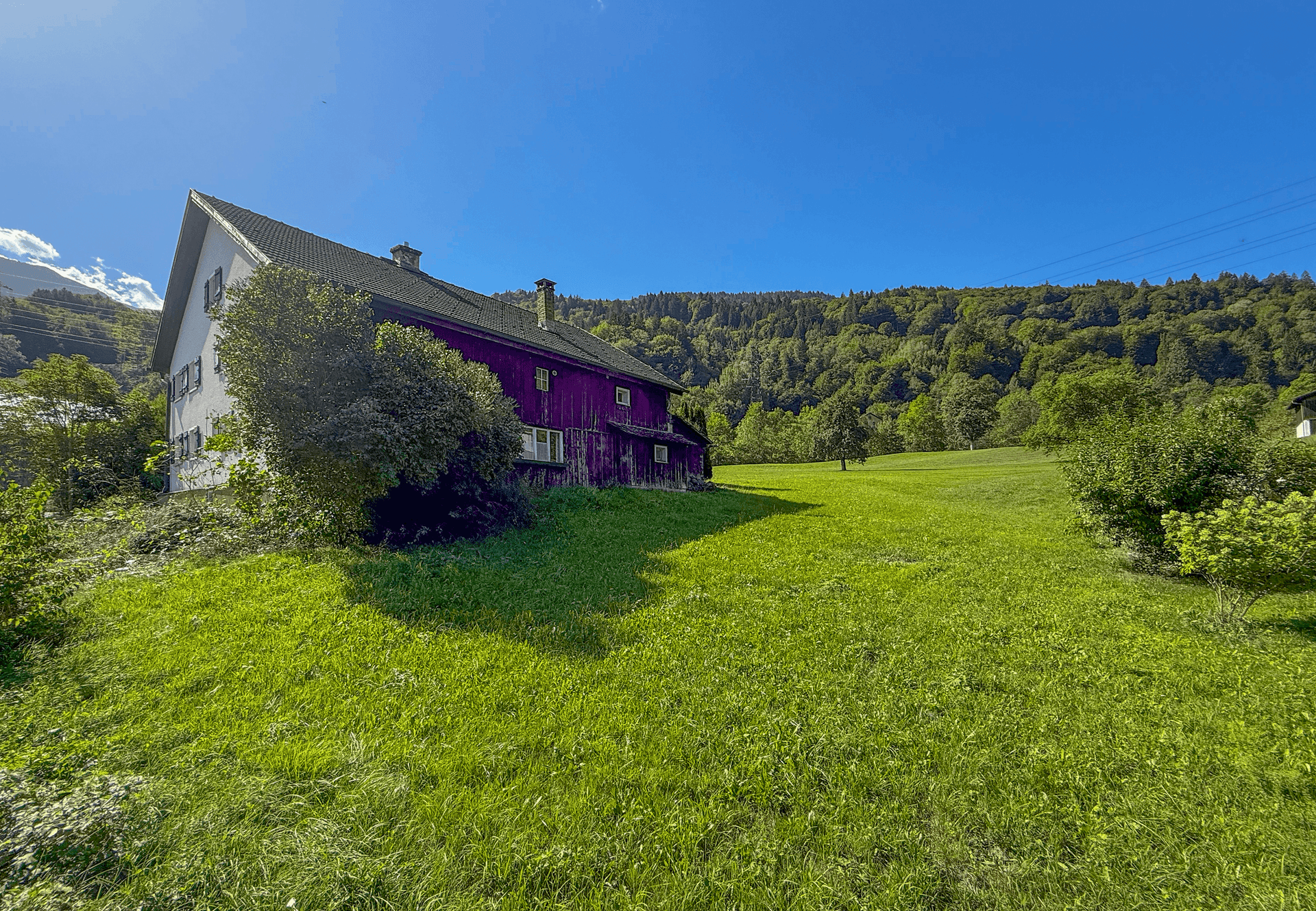 Seitenansicht des Hauses mit weitläufiger grüner Wiese und bewaldeten Hügeln im Hintergrund unter blauem Himmel.
