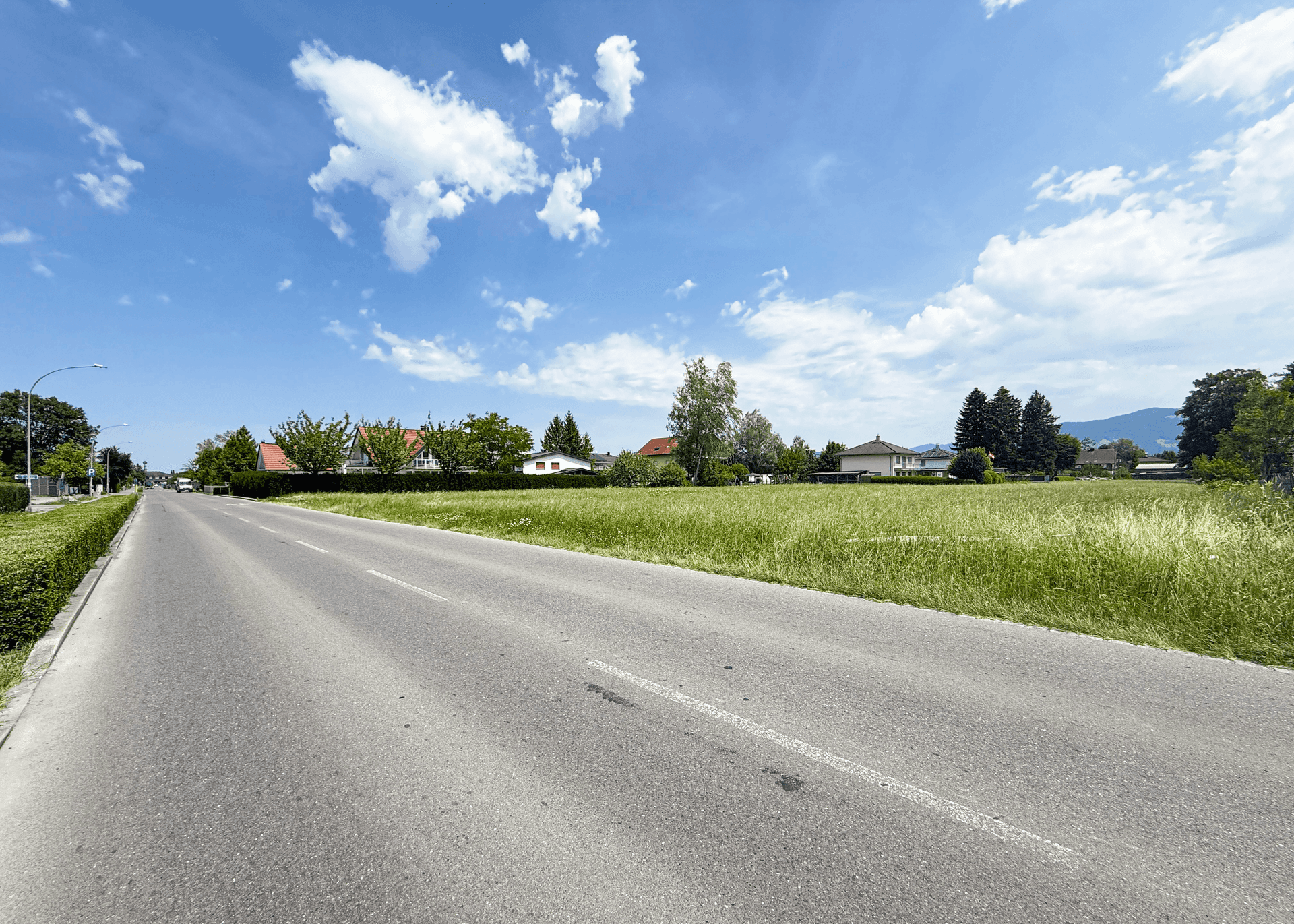 Blick entlang der Straße mit angrenzenden Grünflächen und Wohnhäusern unter einem klaren Himmel.