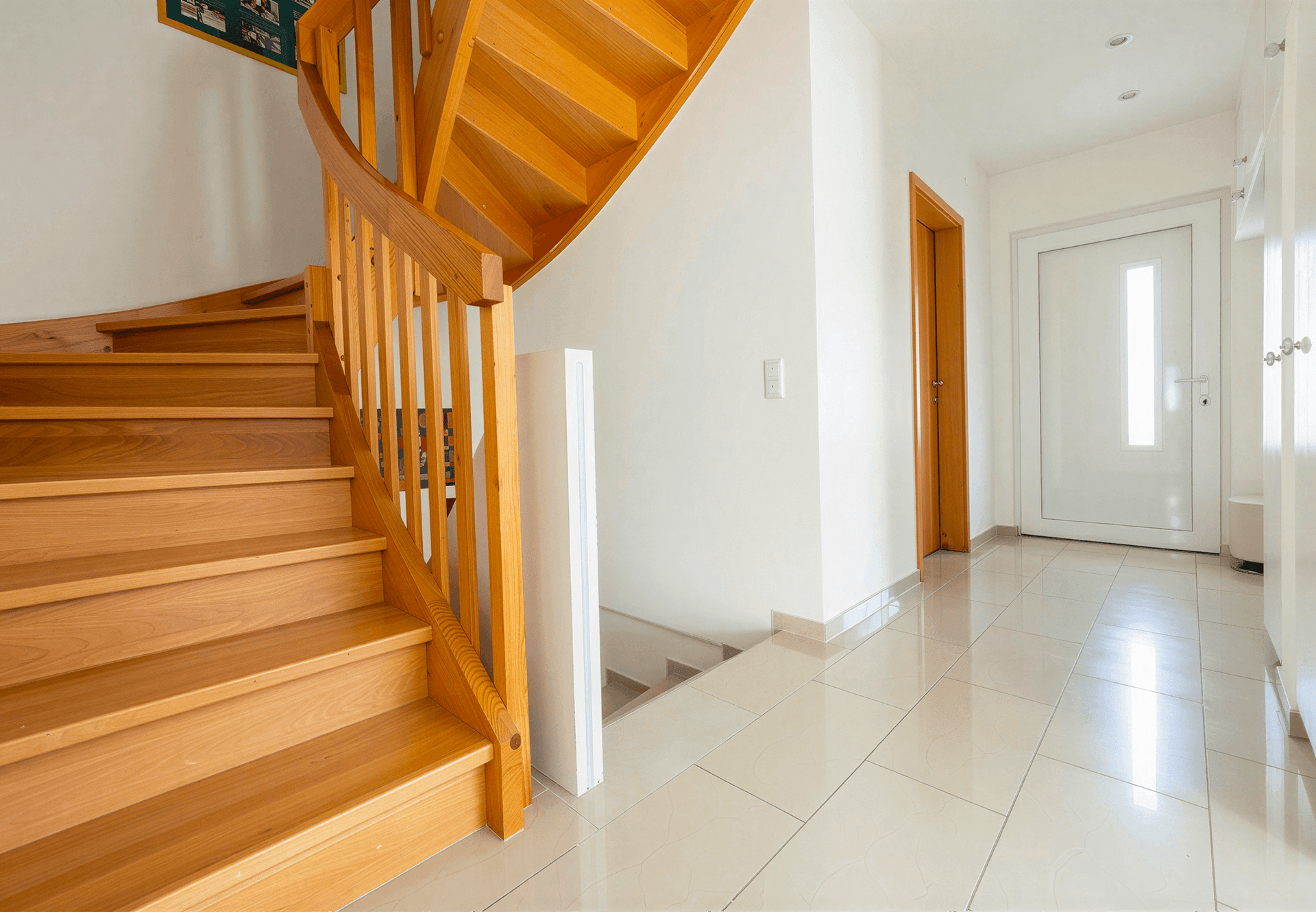 Heller Flur mit eleganter Holztreppe, glänzendem Fliesenboden und weißer Eingangstür, sehr einladend.