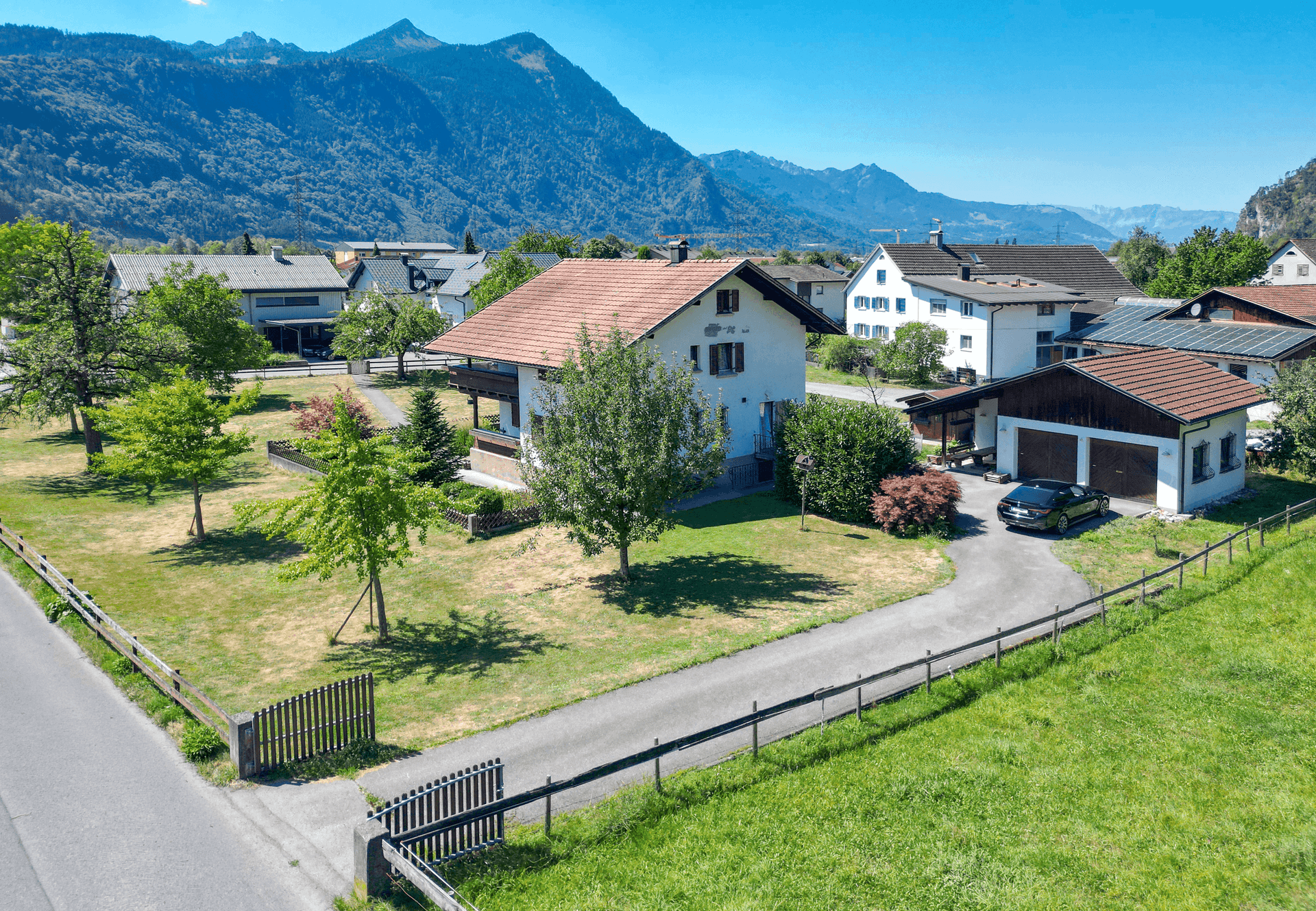 Panoramablick auf das Einfamilienhaus mit großem Garten, Garage und majestätischem Bergpanorama im Hintergrund.