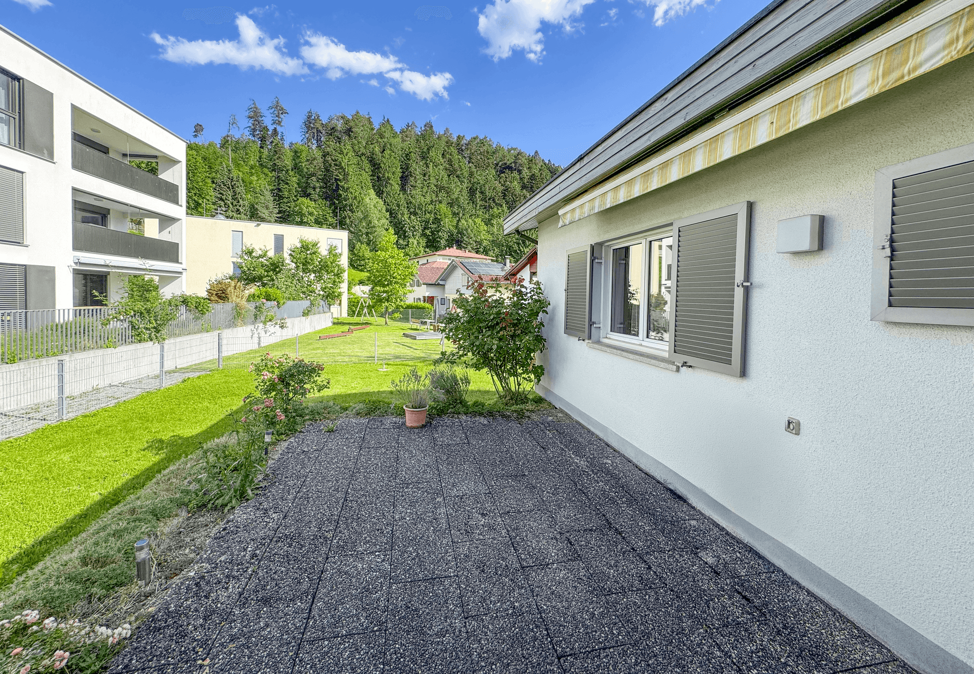 Gepflasterte Terrasse neben dem Haus mit angrenzendem Rasen und Blick auf die umliegende Natur.