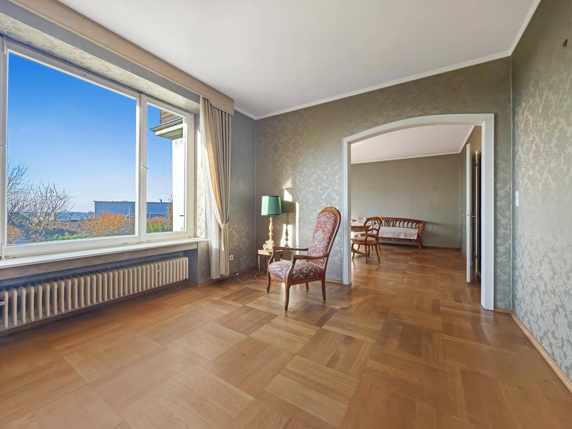 Helles Wohnzimmer mit Parkettboden, großen Fenstern und Zugang zu einem weiteren Raum.