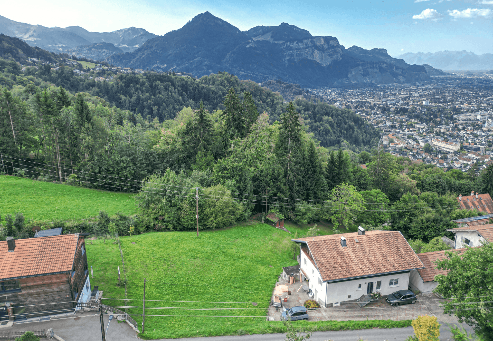 Erhöhte Ansicht von Häusern, grünen Hügeln und einer fernen Stadtkulisse mit majestätischen Bergen im Hintergrund.