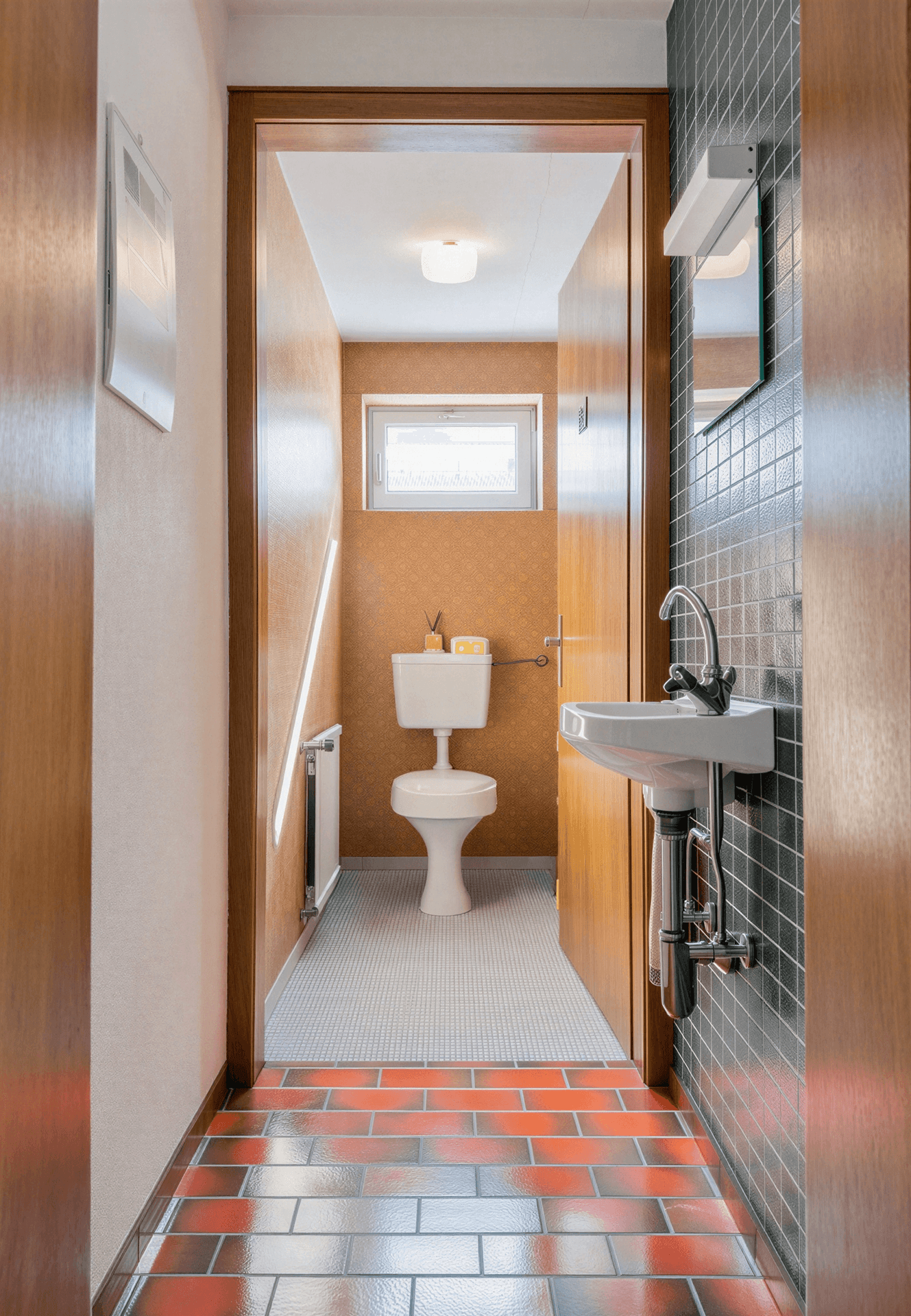 Kleines Gäste-WC mit Waschbecken, Toilette und Fenster, zugänglich über den gefliesten Flur.