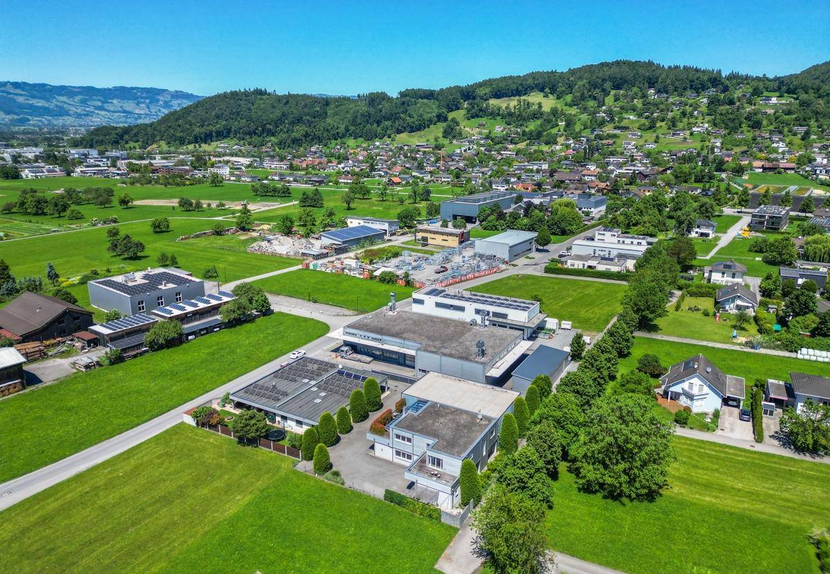 Luftaufnahme eines Gewerbegebiets mit mehreren Gebäuden, umgeben von grünen Wiesen und Bergen.
