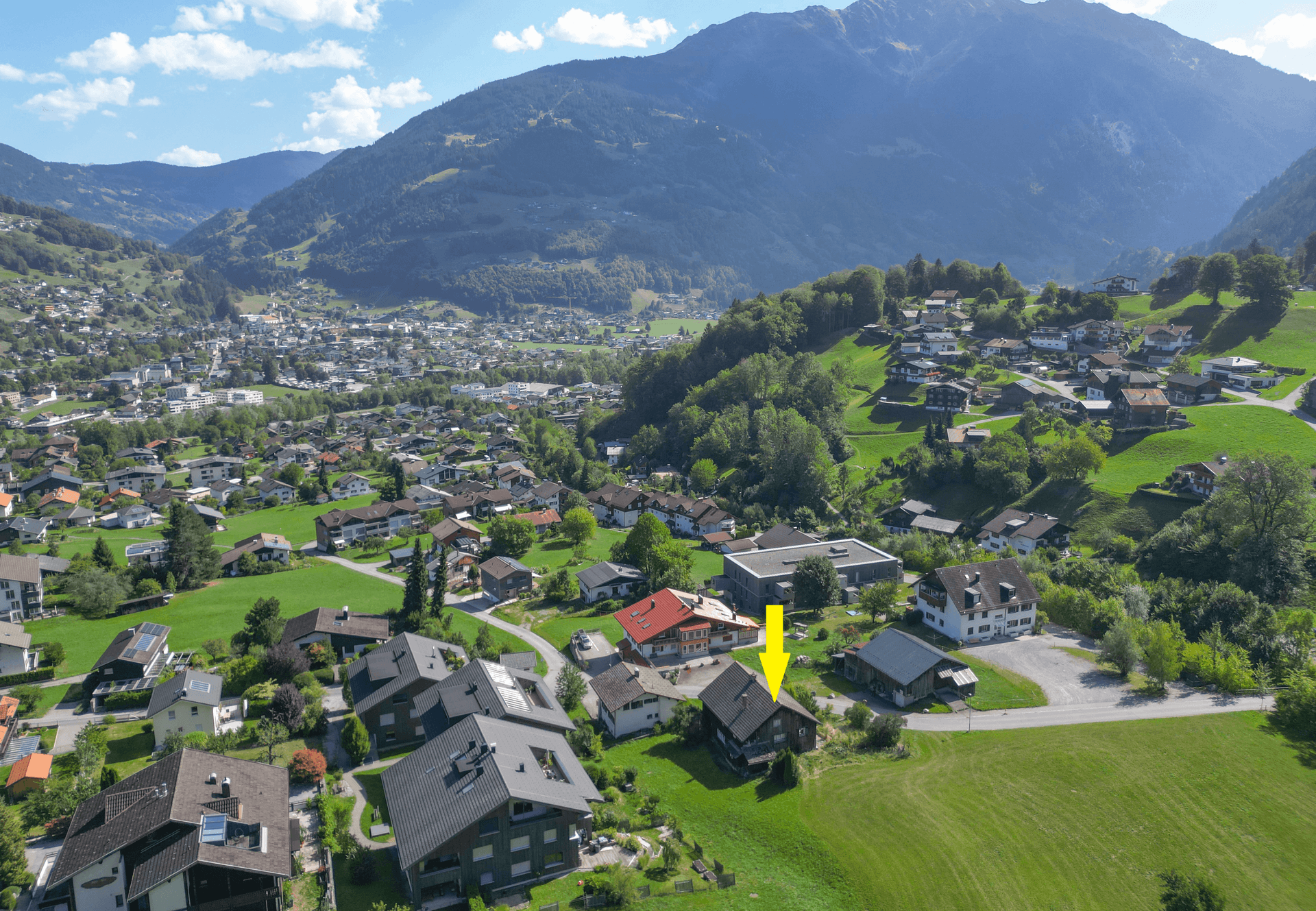 Weitläufige Luftaufnahme des Tals mit dem Dorf und dem markierten Objekt, umgeben von bewaldeten Bergen.
