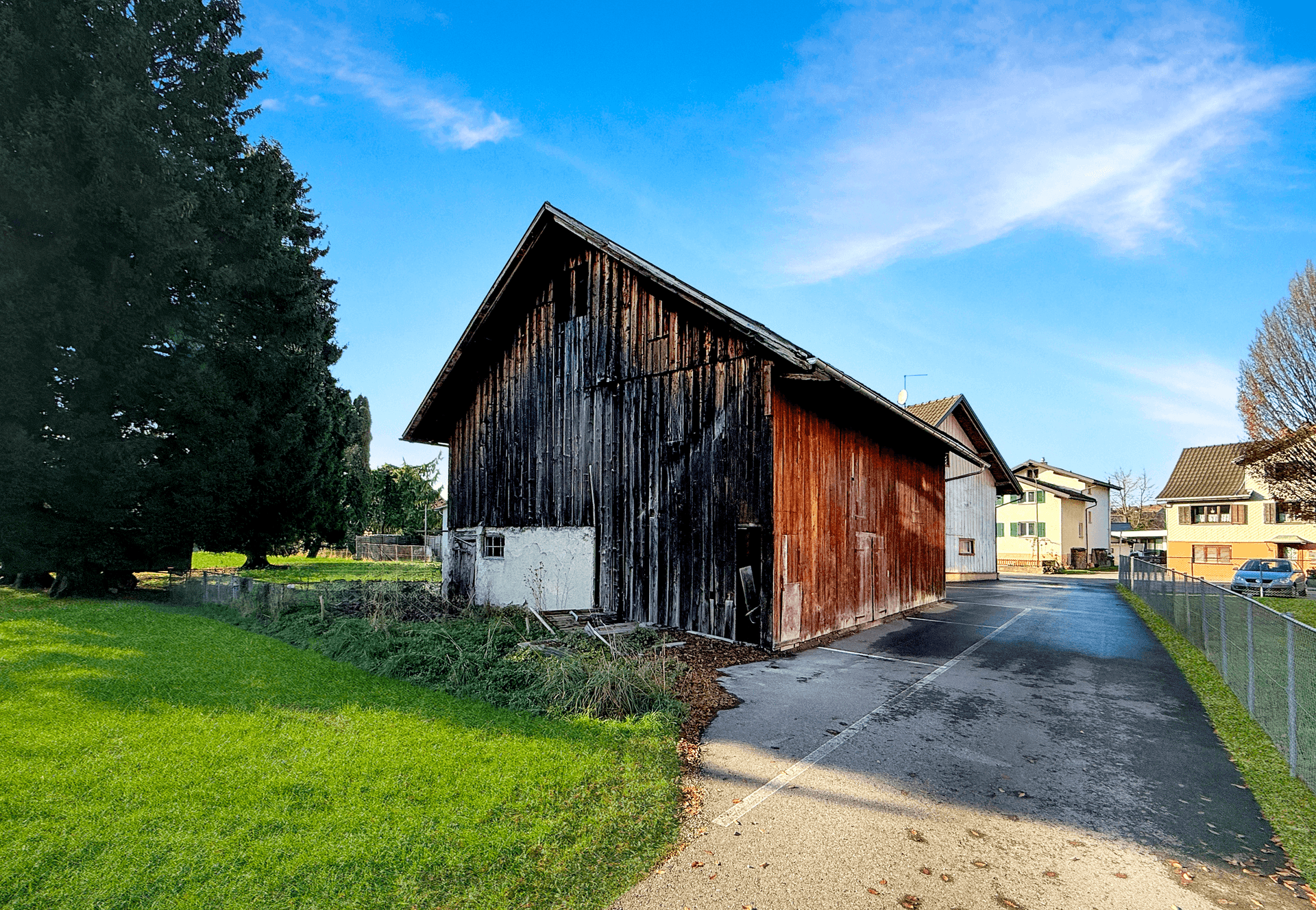 Langes, rustikales Holzgebäude mit verwitterter Fassade und einem asphaltierten Weg daneben.