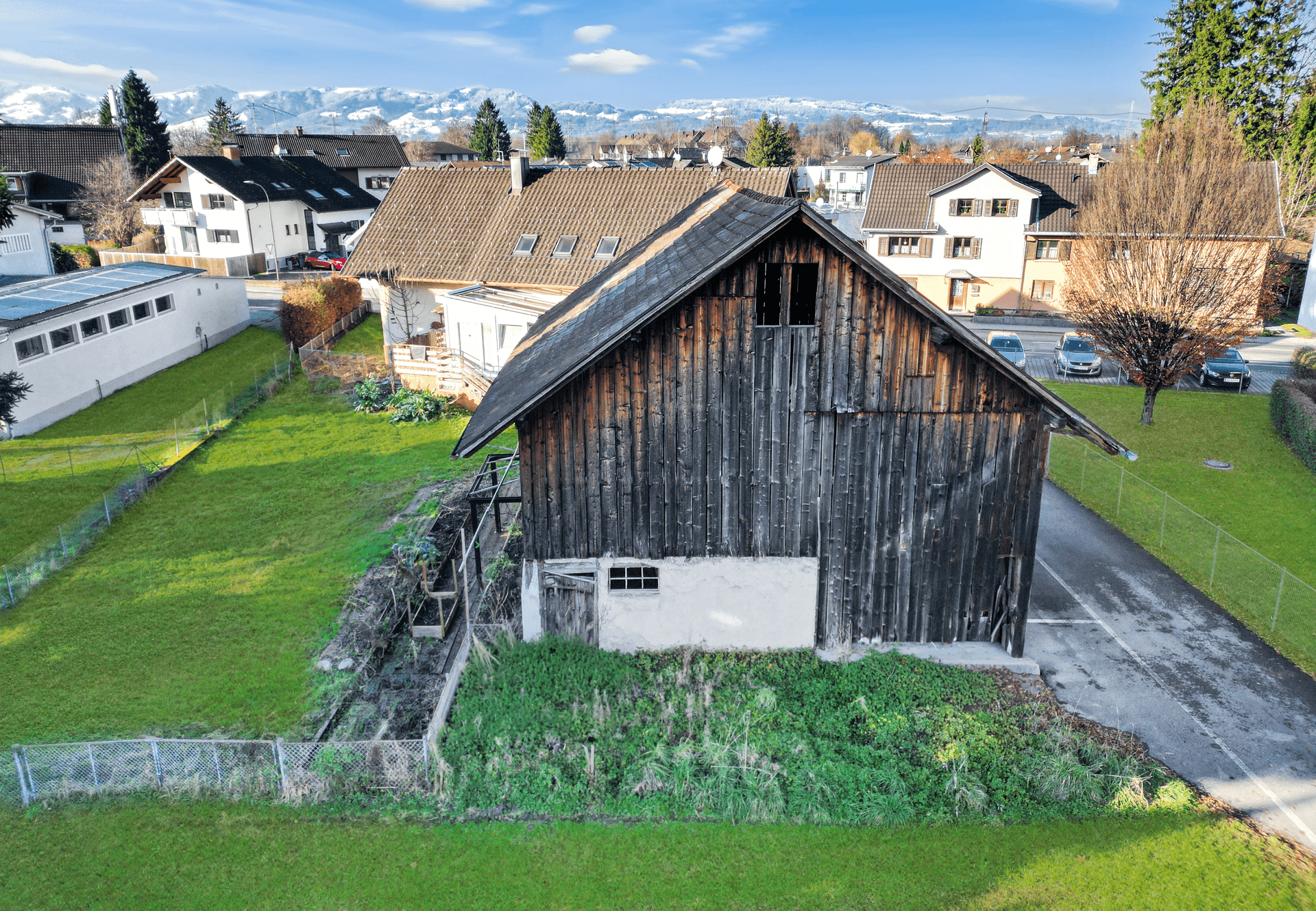 Rustikales Holzgebäude mit angrenzendem Wohnhaus und grünem Garten vor einer Bergkulisse.