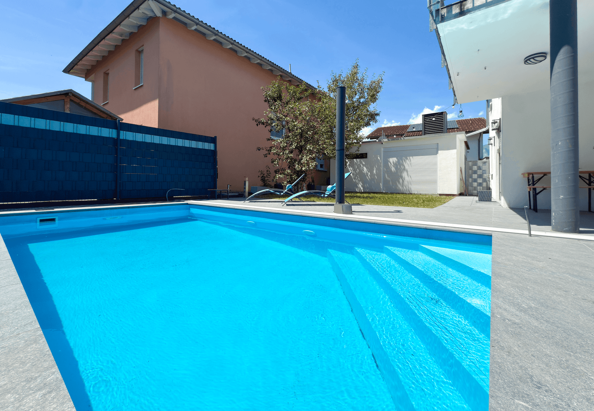 Blauer Pool mit Treppeneinstieg und umliegender Terrasse, umgeben von Häusern.