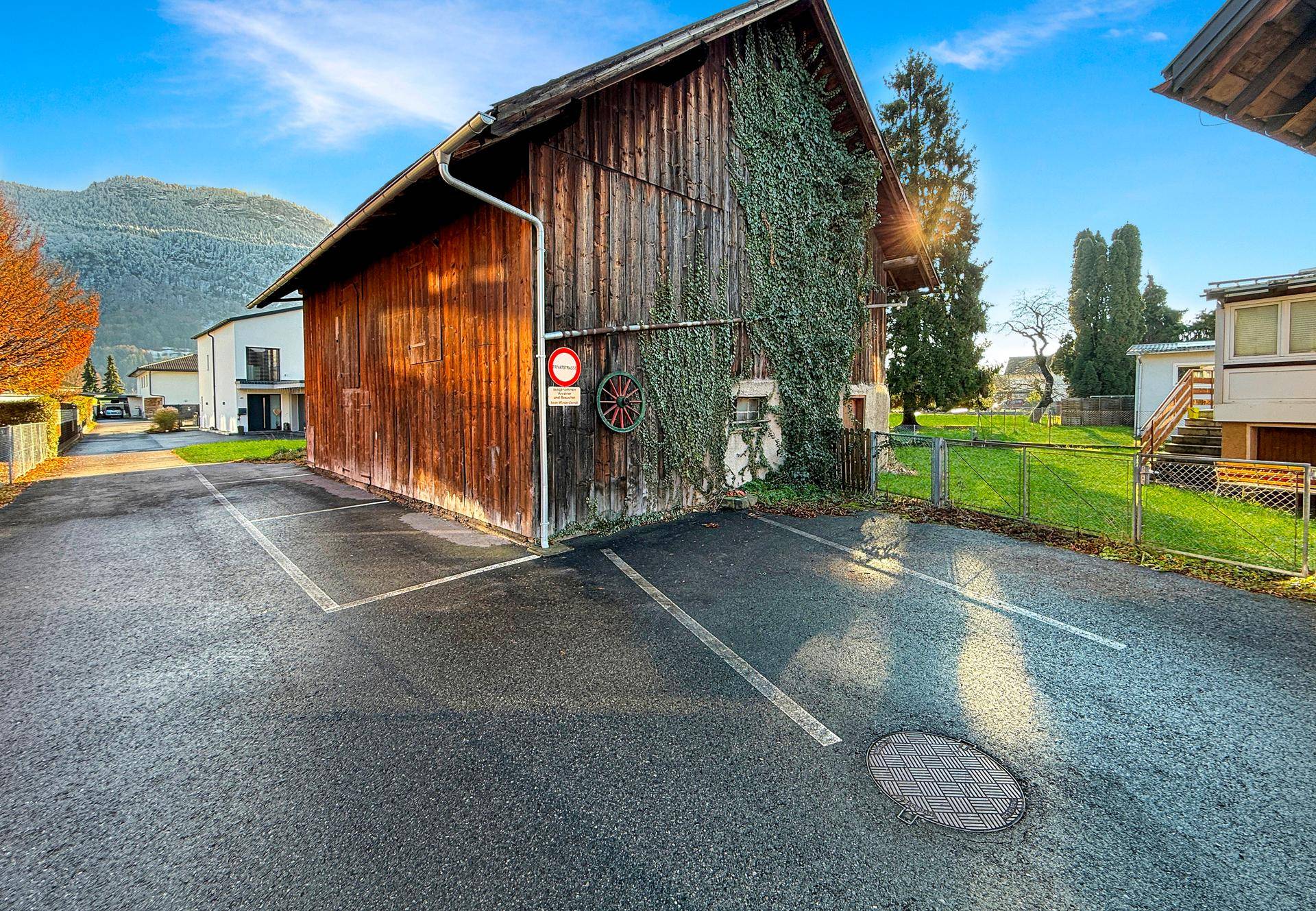 Altes Holzgebäude mit Efeubewuchs und asphaltiertem Parkplatz vor einer bergigen Landschaft.