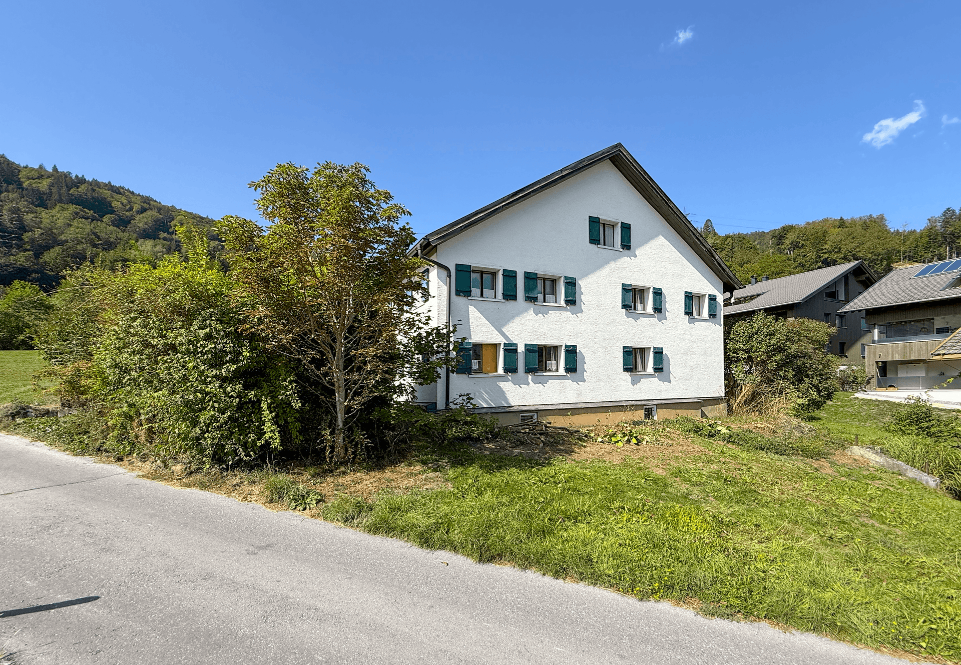 Seitenansicht des Hauses mit weißer Fassade, grünen Fensterläden und einer ruhigen Landstraße im Vordergrund.