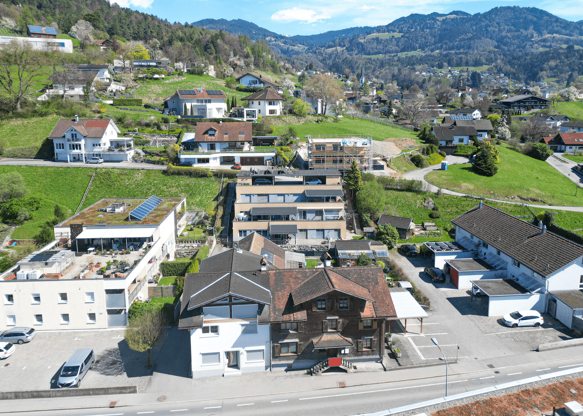 Luftaufnahme einer Wohnsiedlung mit Neubauten, grünen Hügeln und Bergen im Hintergrund.