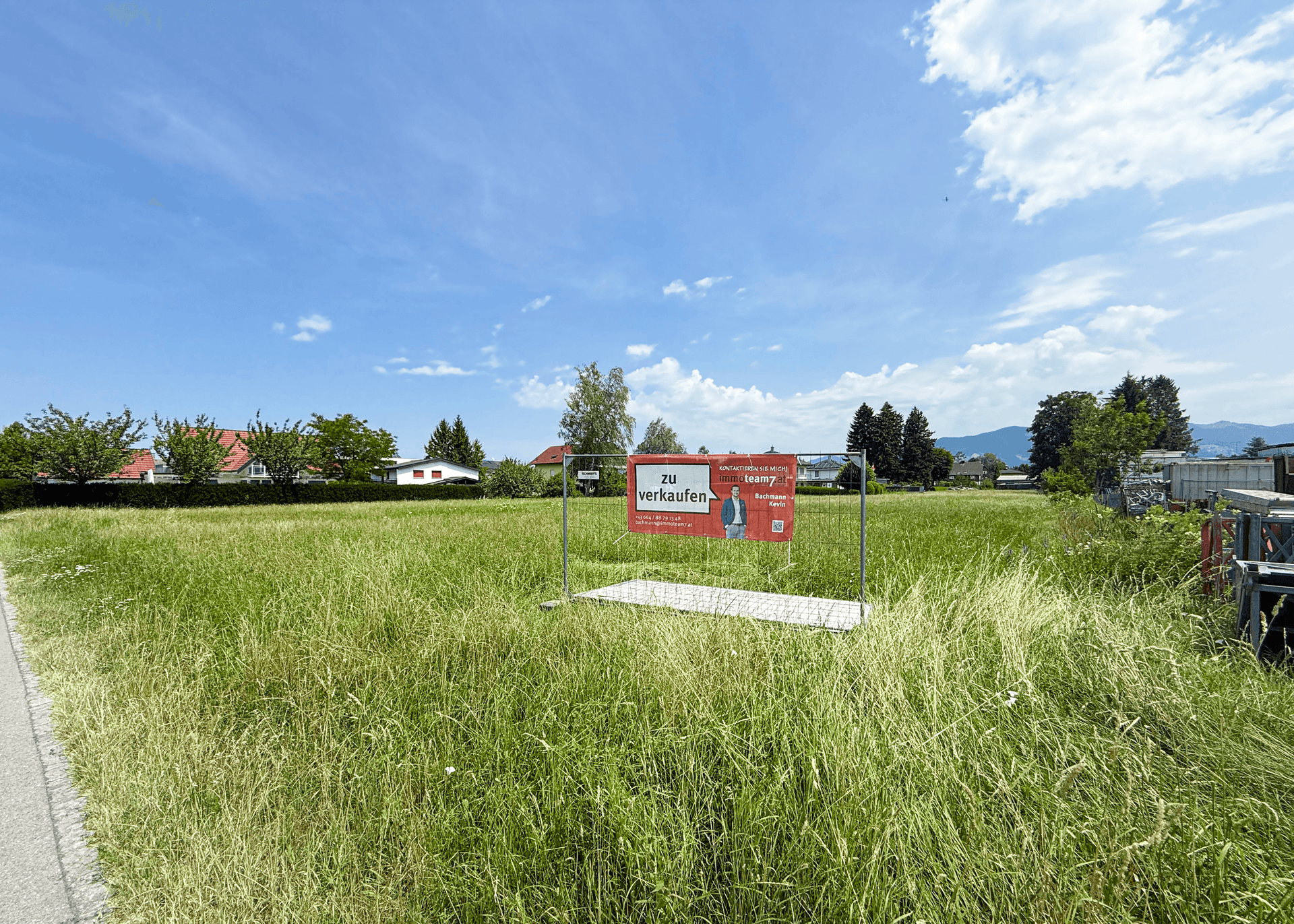 Grünes Baugrundstück mit einem 'Zu verkaufen'-Schild unter einem weiten blauen Himmel mit Wolken.