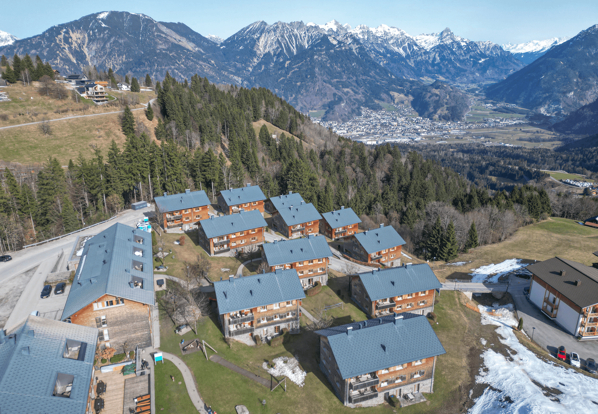 Panoramablick auf die Ferienanlage mit Holzhäusern, eingebettet in ein weites Tal mit majestätischen Bergen.