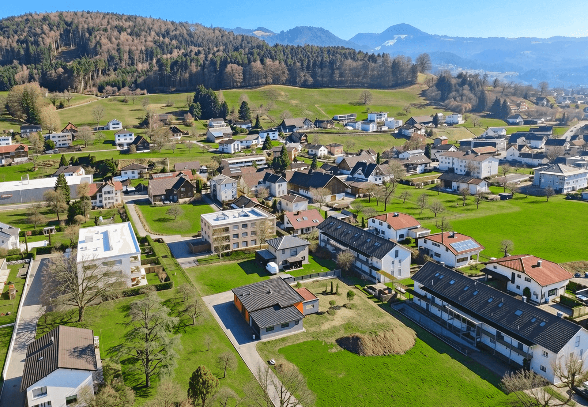 Luftbild einer Wohngegend mit viel Grün, umgeben von bewaldeten Hügeln und Bergen.