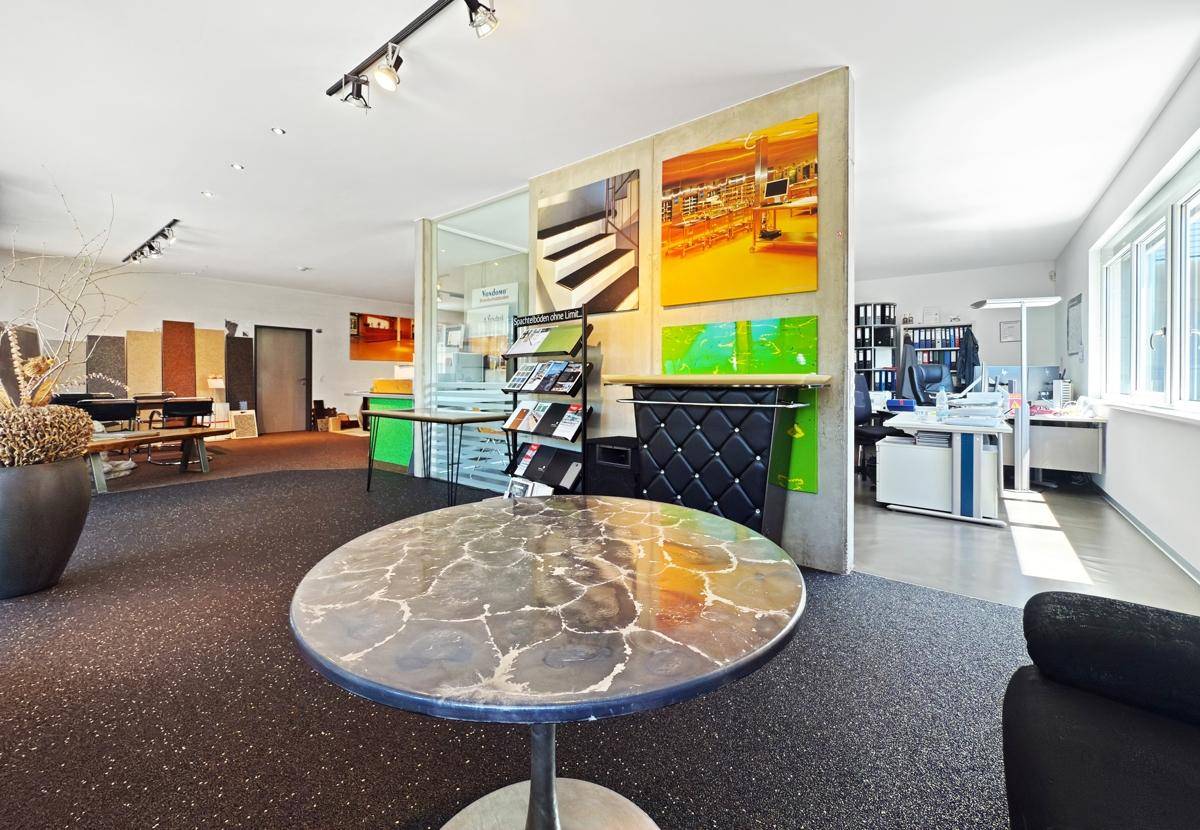Geräumiger Ausstellungsraum mit verschiedenen Bodenbelägen, einem runden Tisch und Bürobereich im Hintergrund.