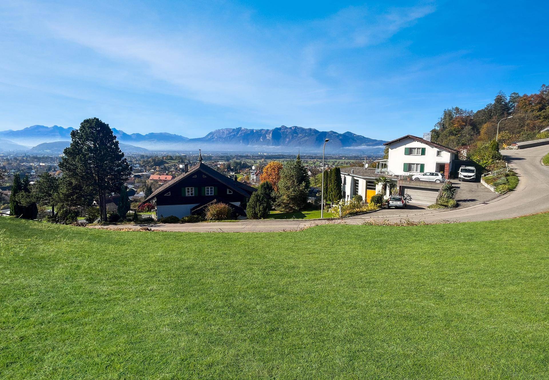 Weitläufige Grünfläche mit Häusern am Hang und beeindruckendem Bergpanorama unter blauem Himmel.