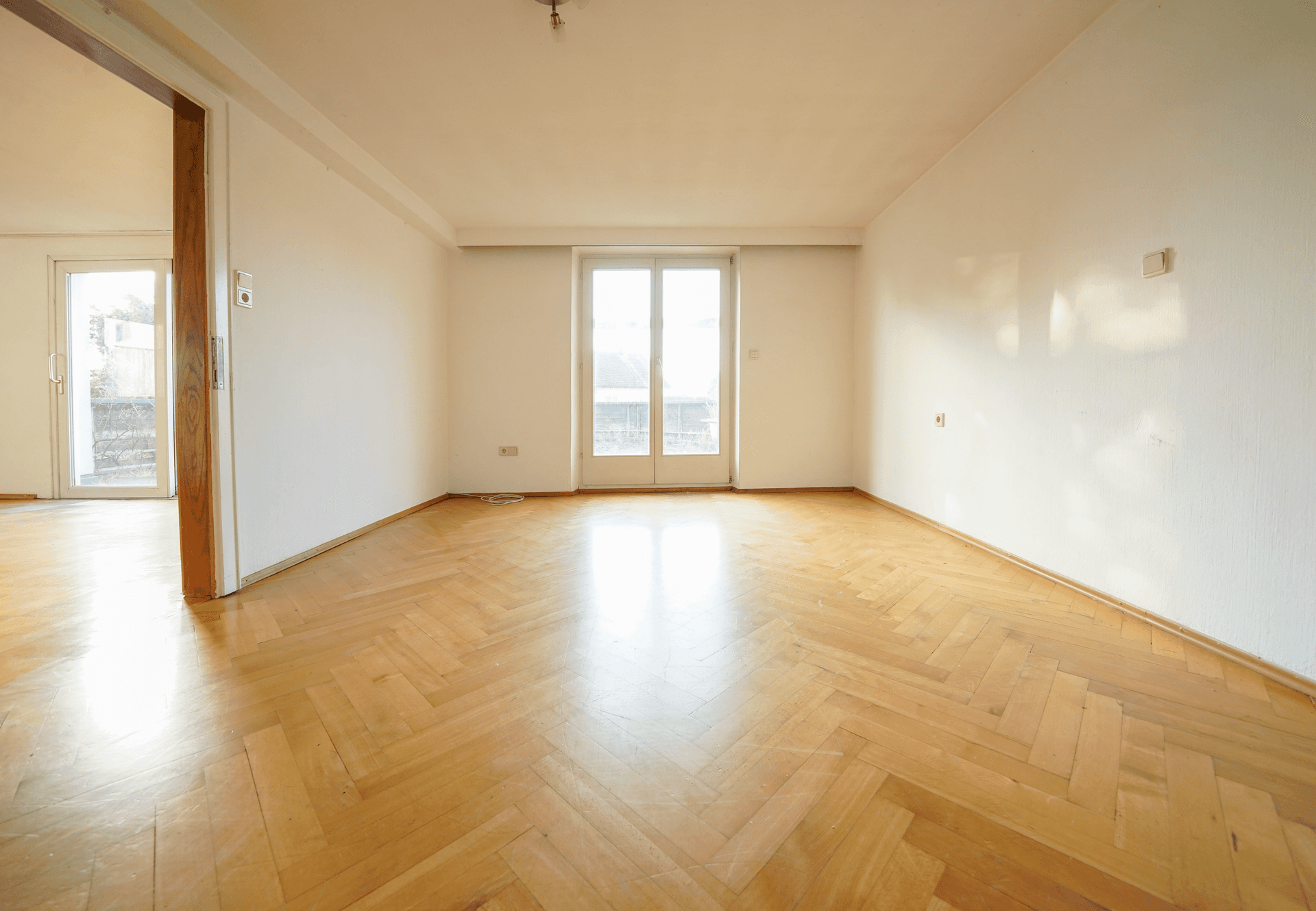Helles, leeres Zimmer mit Fischgrätparkettboden und Zugang zu einem Balkon oder einer Terrasse.