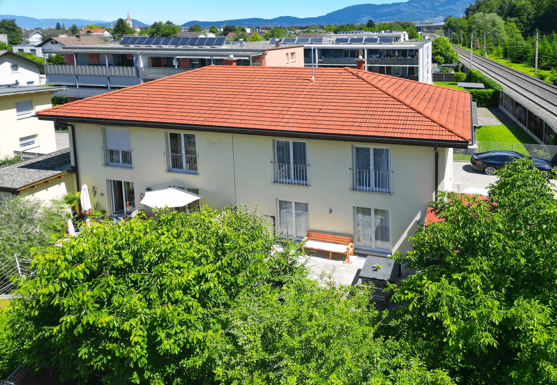 Luftaufnahme des Hauses mit rotem Dach, Balkonen und umliegender Wohnbebauung, eingebettet in Grün.