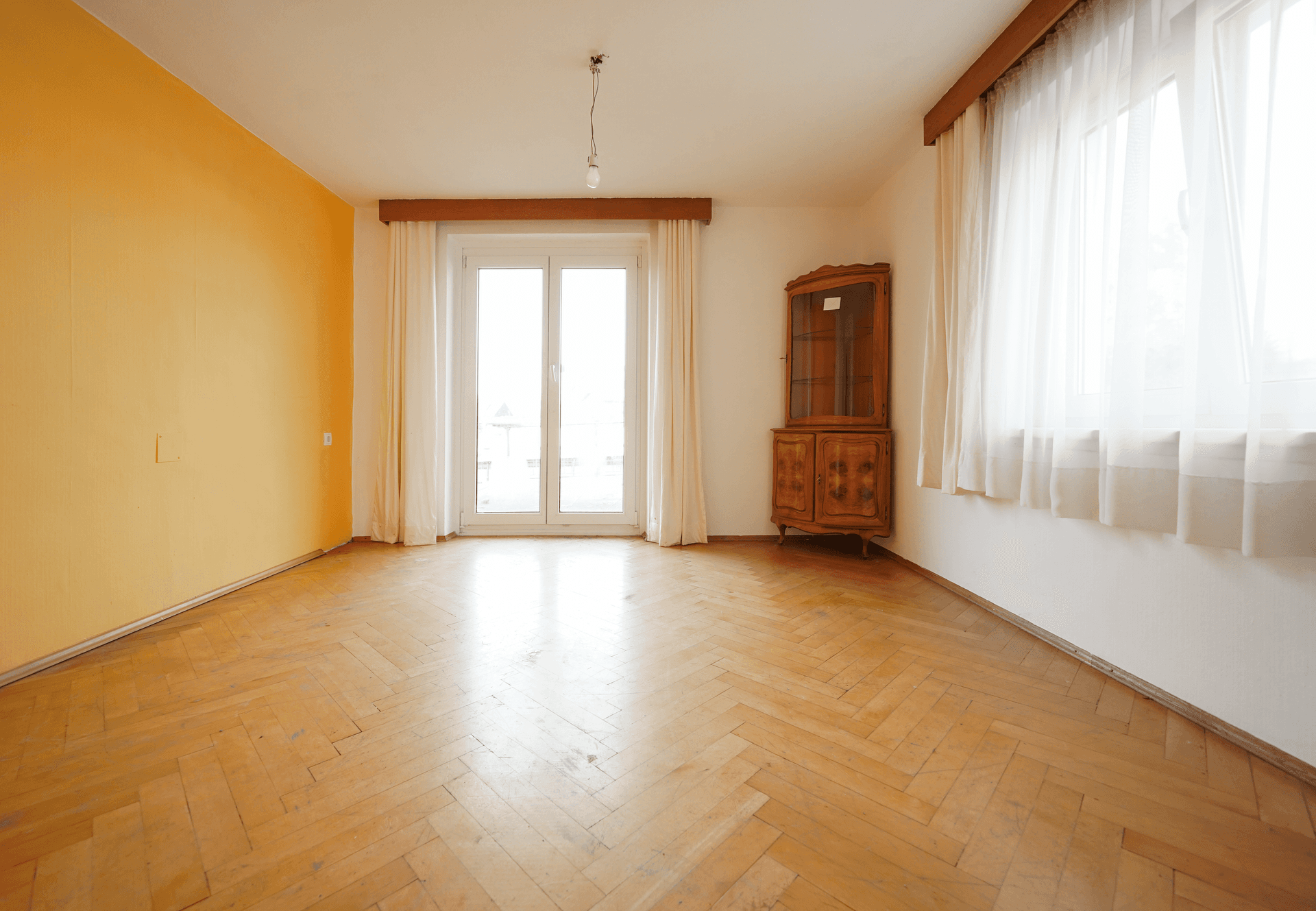 Leeres Zimmer mit Fischgrätparkett, einem Fenster und einem antiken Holzschrank.