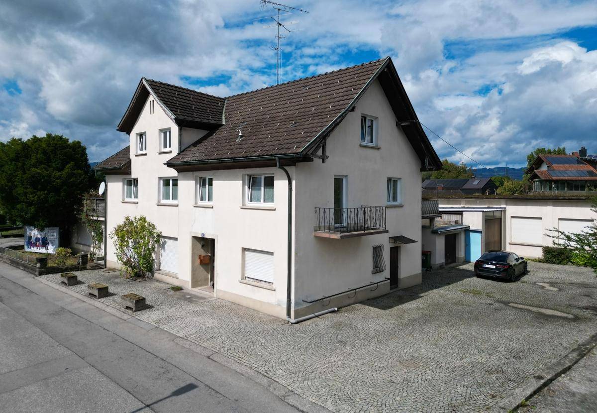 Ansicht des Wohnhauses mit weißer Fassade, kleinem Balkon und gepflastertem Vorplatz.