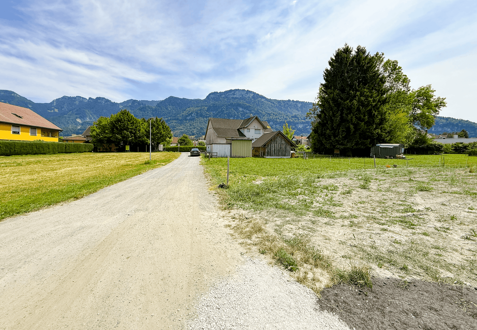 Unbefestigter Weg führt durch grüne Wiesen zu einem Haus mit Bergkulisse im Hintergrund.
