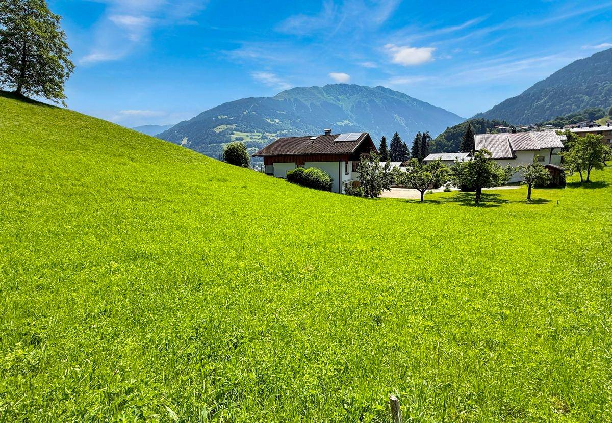 Einfamilienhaus umgeben von grünen Wiesen mit Bergblick unter strahlend blauem Himmel.