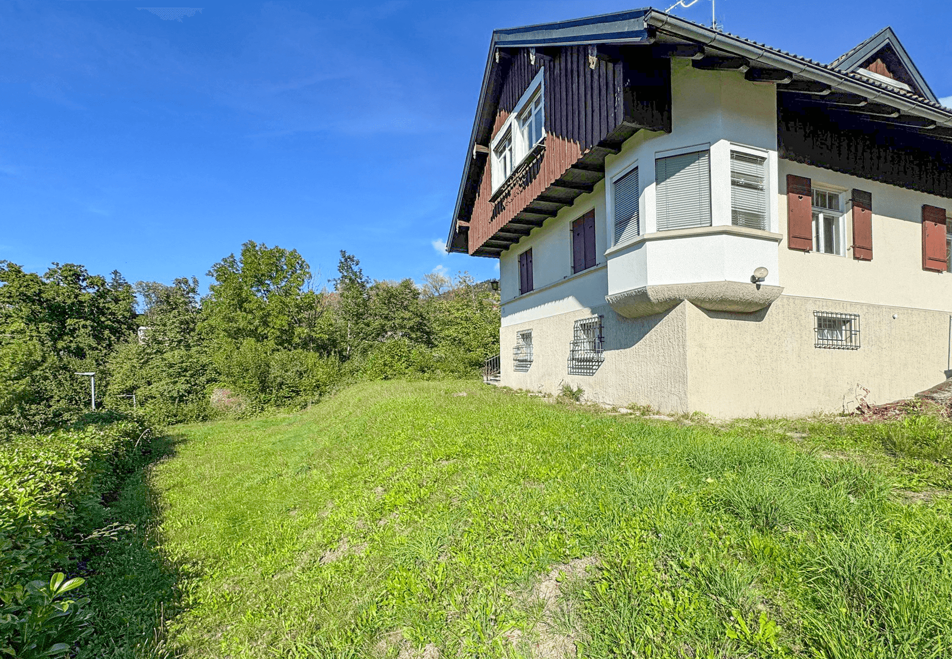 Seitenansicht des Hauses mit Erker und grüner Hanglage unter strahlend blauem Himmel.