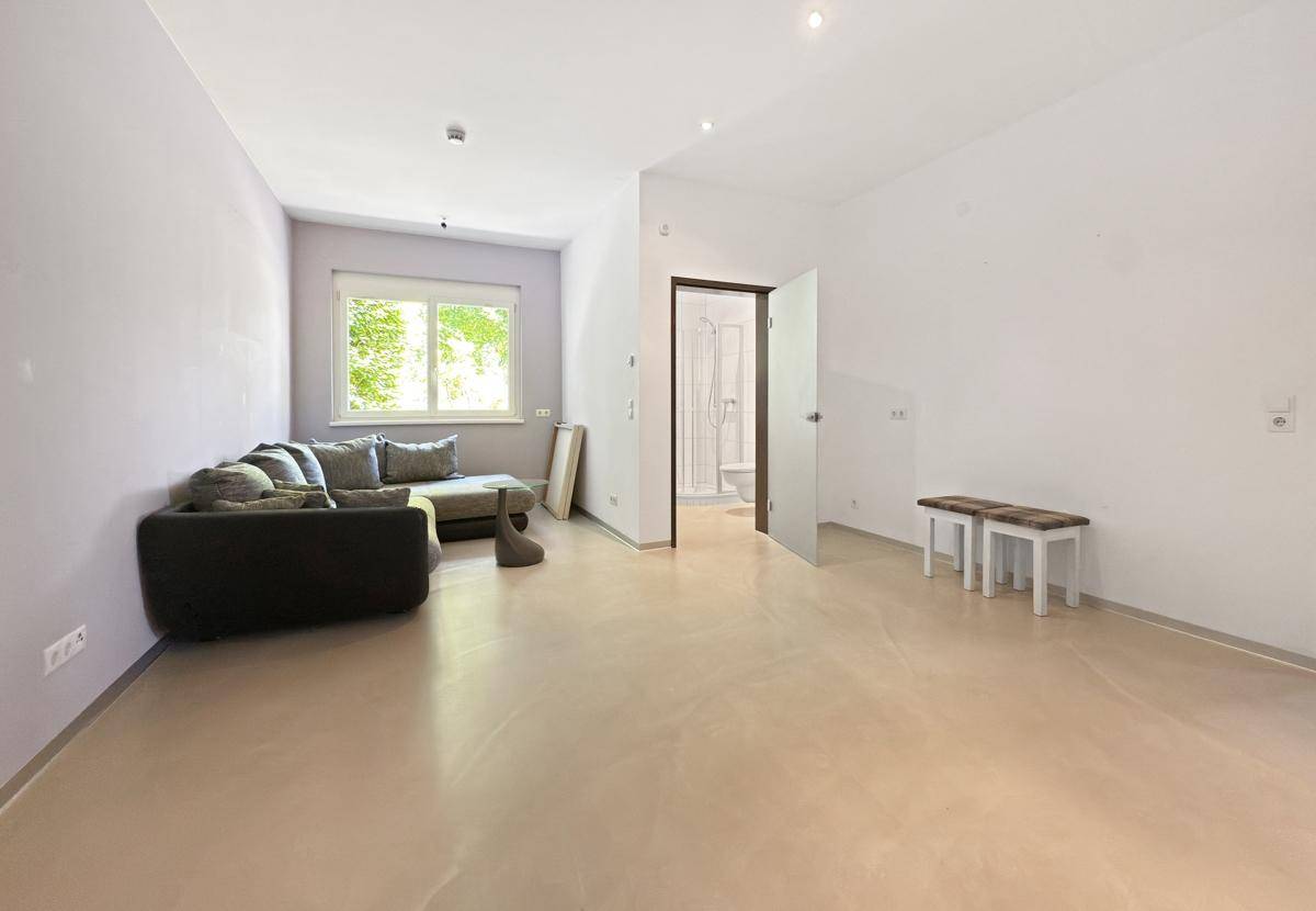 Helles Wohnzimmer mit Sofa, hellem Boden und Zugang zu einem angrenzenden Badezimmer mit Dusche.