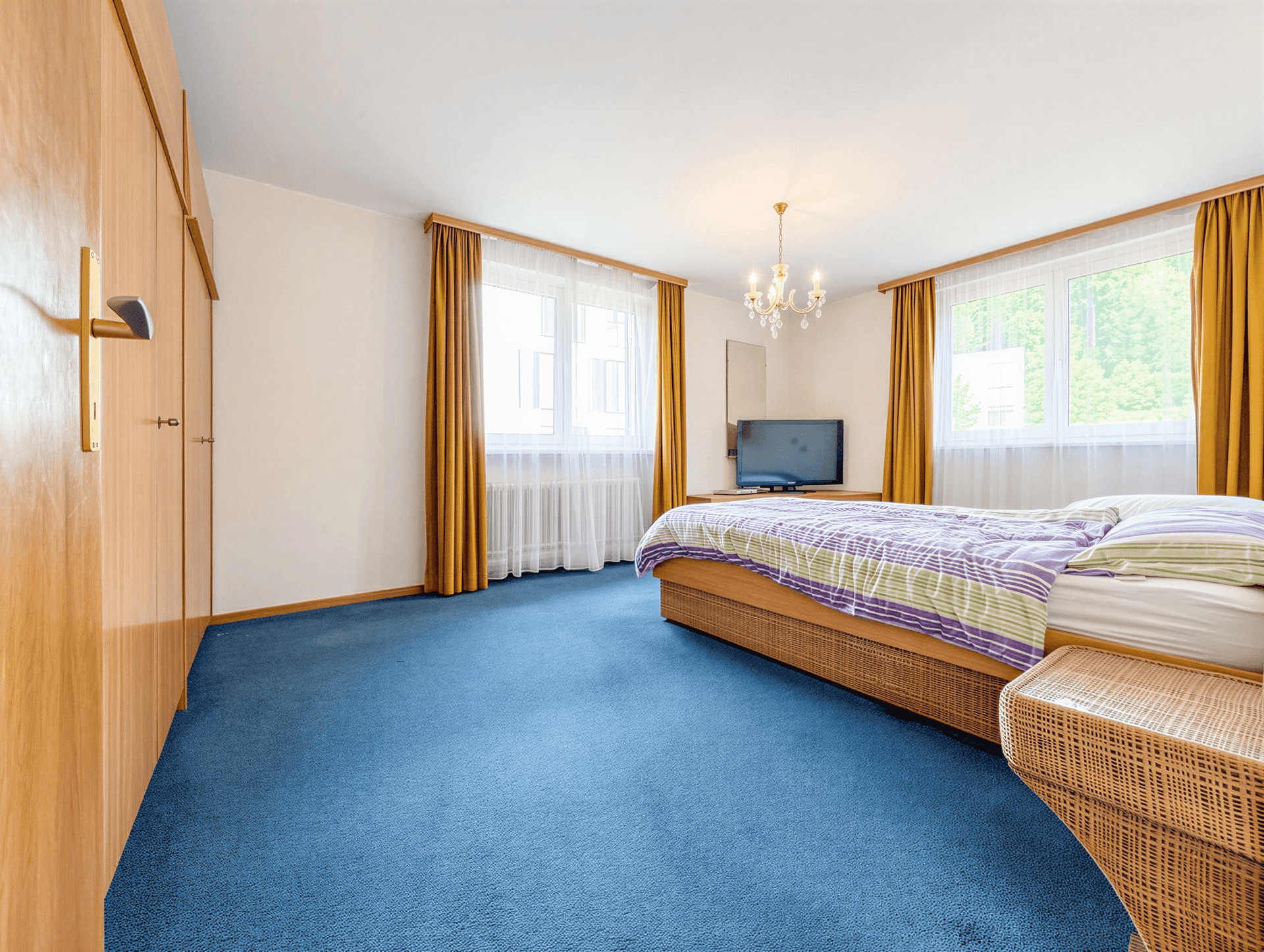 Geräumiges Schlafzimmer mit blauem Teppichboden, großem Bett und viel Tageslicht durch zwei Fenster.