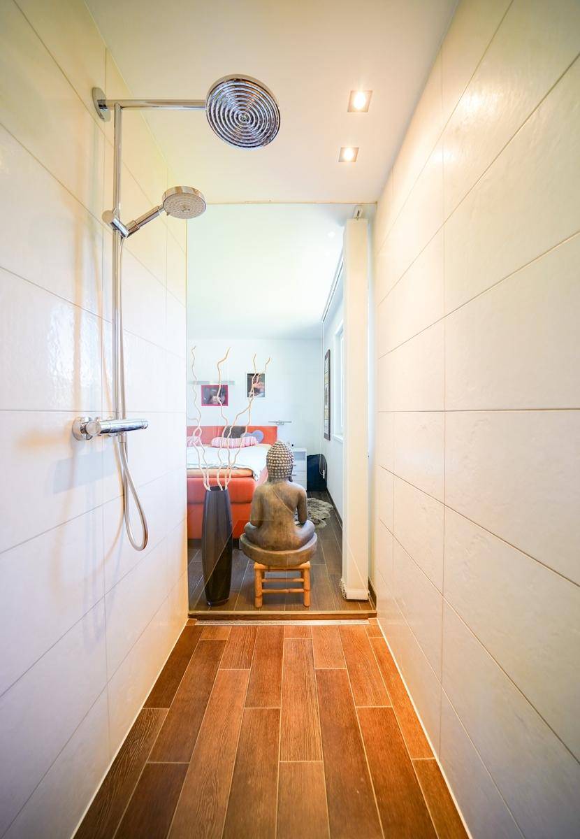 Ebenerdige Dusche mit Holzbodenoptik-Fliesen und Blick in den angrenzenden Raum.