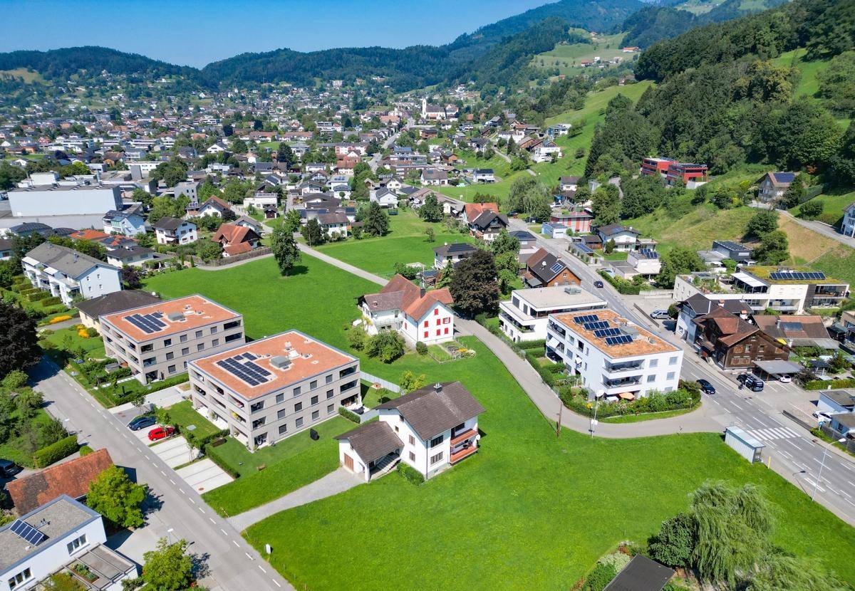 Luftaufnahme einer Wohnsiedlung mit Häusern, Grünflächen und modernen Gebäuden mit Photovoltaikanlagen vor Bergkulisse.