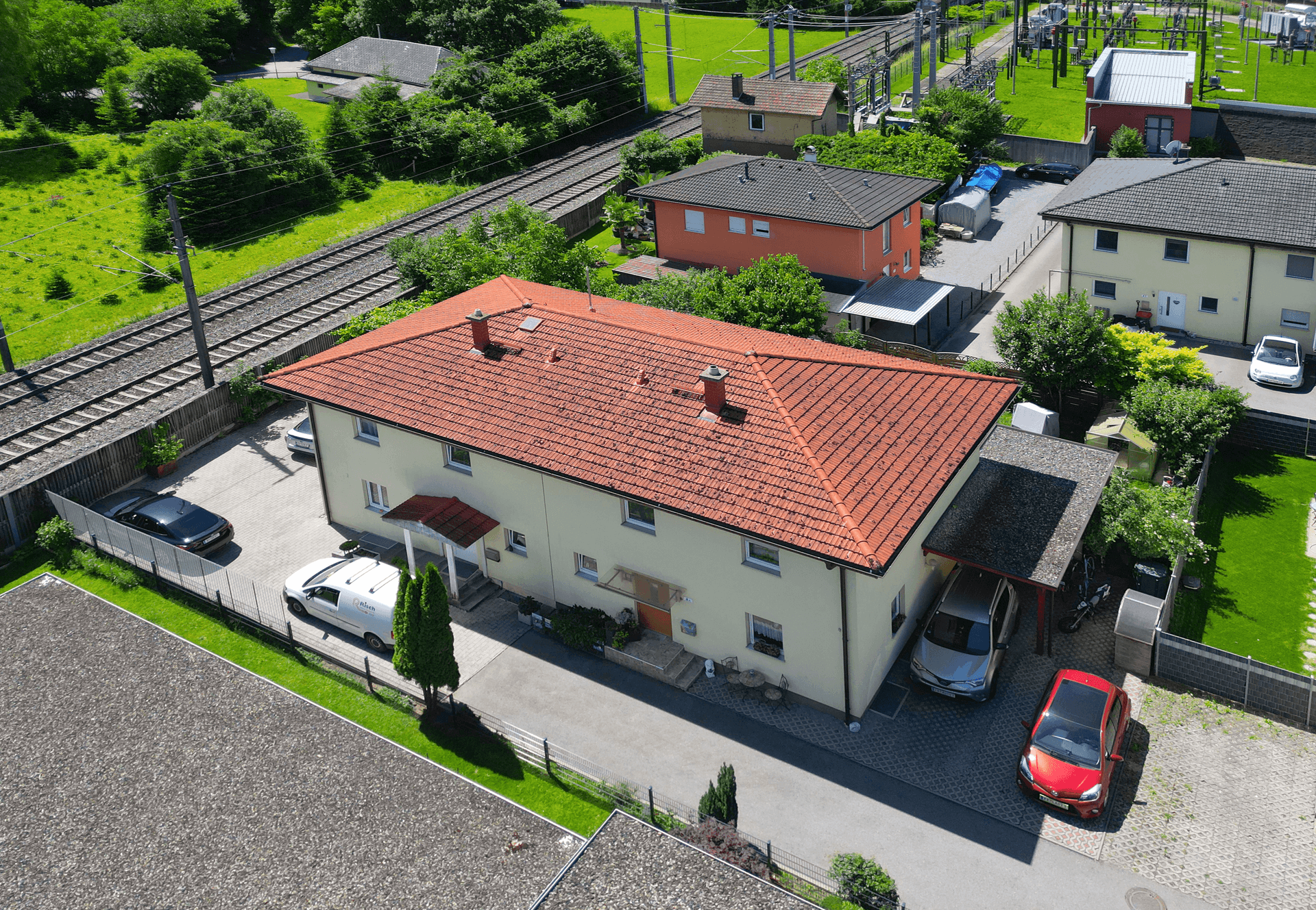 Luftaufnahme des Wohnhauses mit rotem Dach, Carport und angrenzenden Bahngleisen, in sonniger Umgebung.