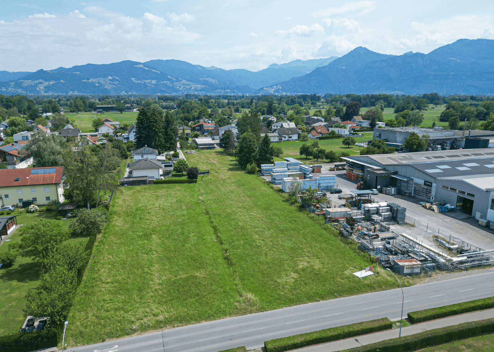 Weitläufige Luftaufnahme des Baugrundstücks mit angrenzenden Gewerbegebäuden und Bergpanorama im Hintergrund.