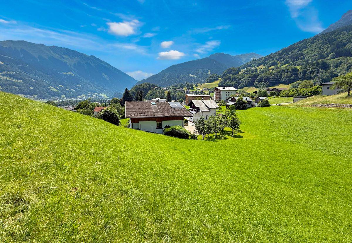 Weitläufige Landschaft mit Häusern in einem grünen Tal, umgeben von hohen Bergen unter blauem Himmel.