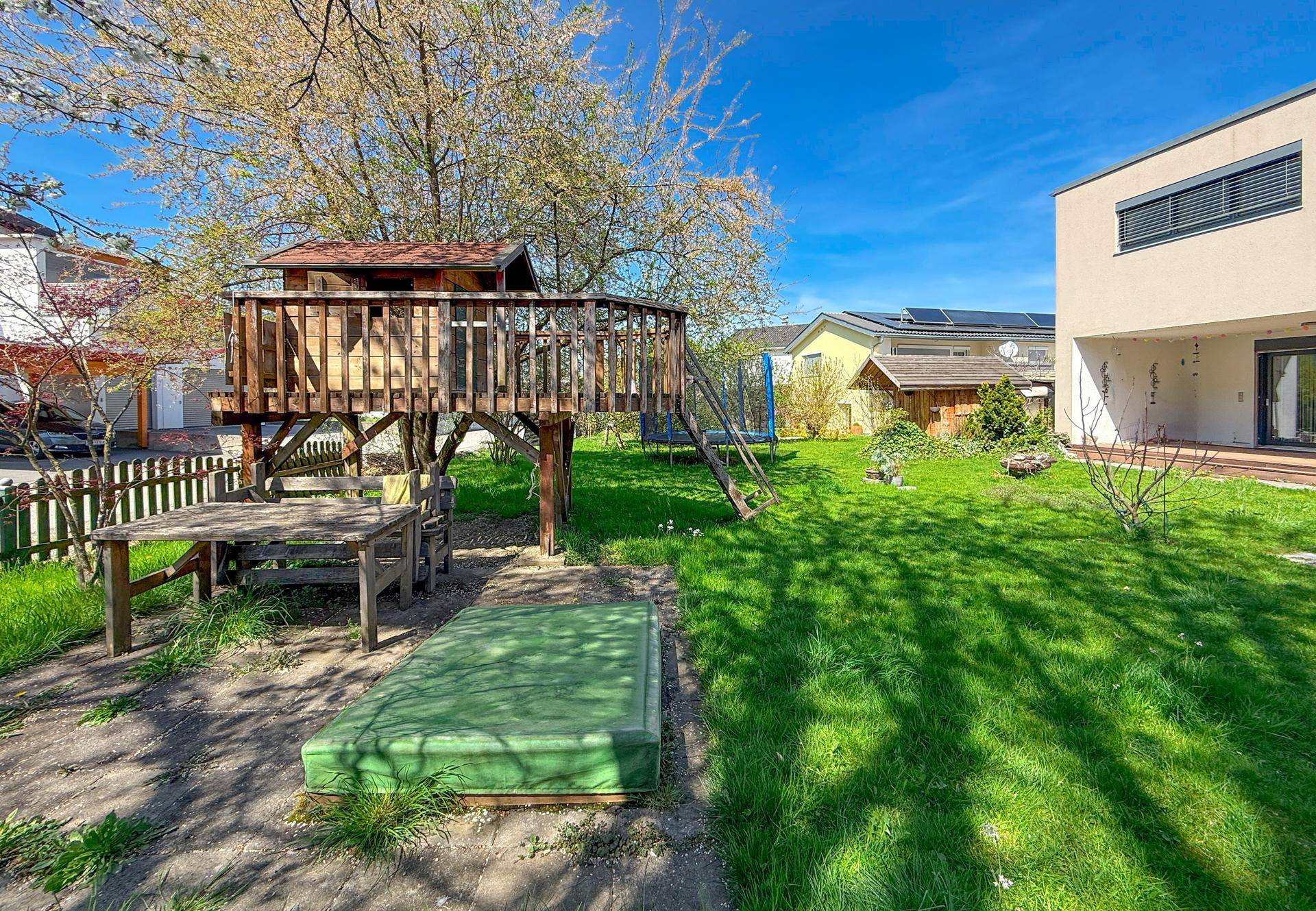 Sonniger Gartenbereich mit Baumhaus, Holztisch und Blick auf das moderne Wohnhaus.