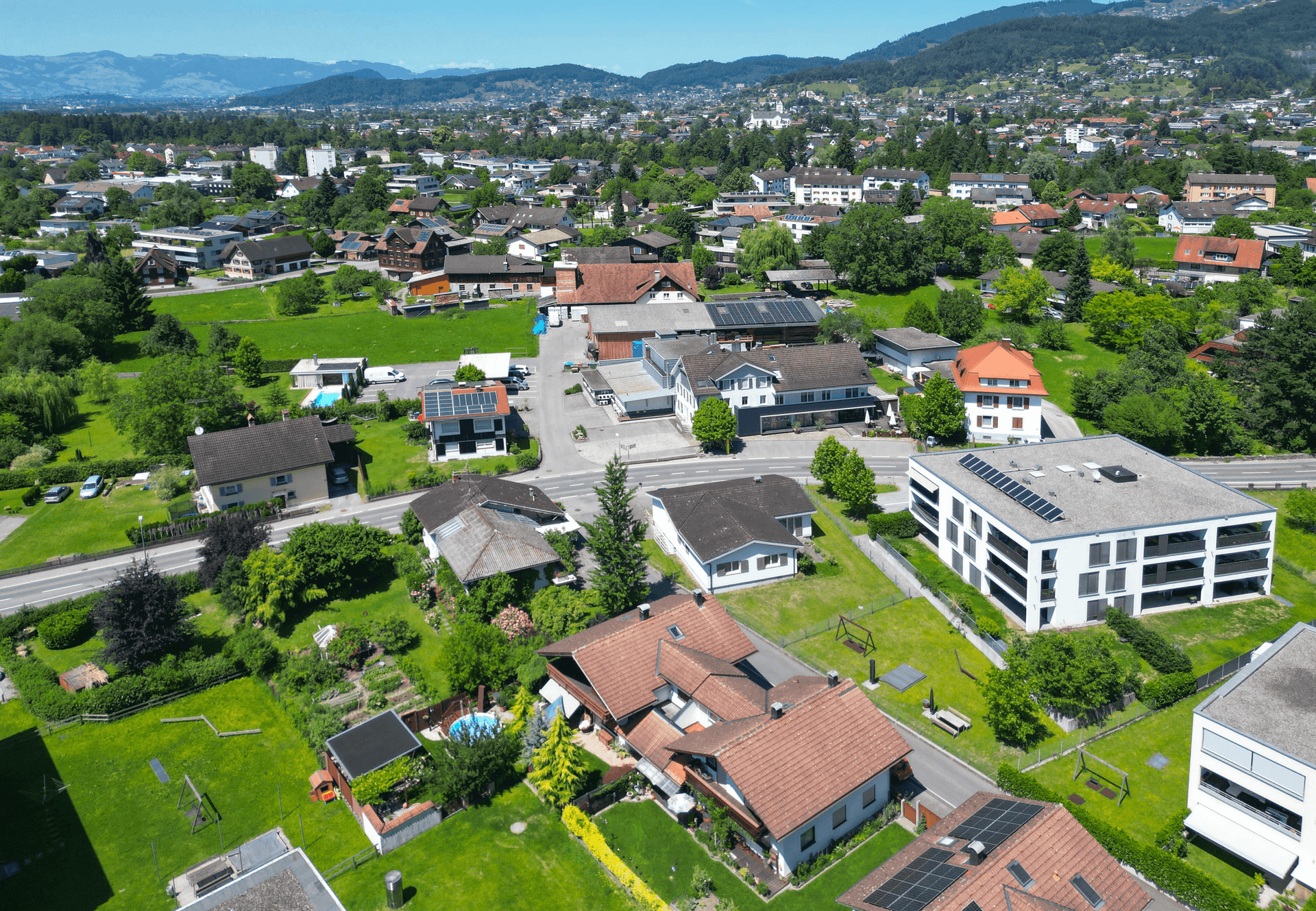 Weitläufige Luftaufnahme der Wohngegend mit vielen Häusern, Grünflächen und Bergen im Hintergrund.