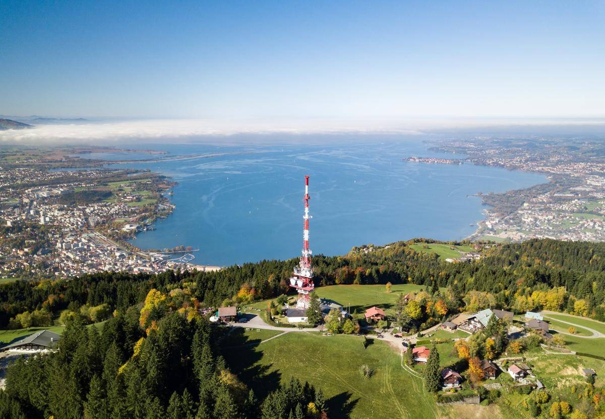 Panoramische Luftaufnahme des Bodensees mit einer Stadt und einem Sendeturm im Vordergrund.