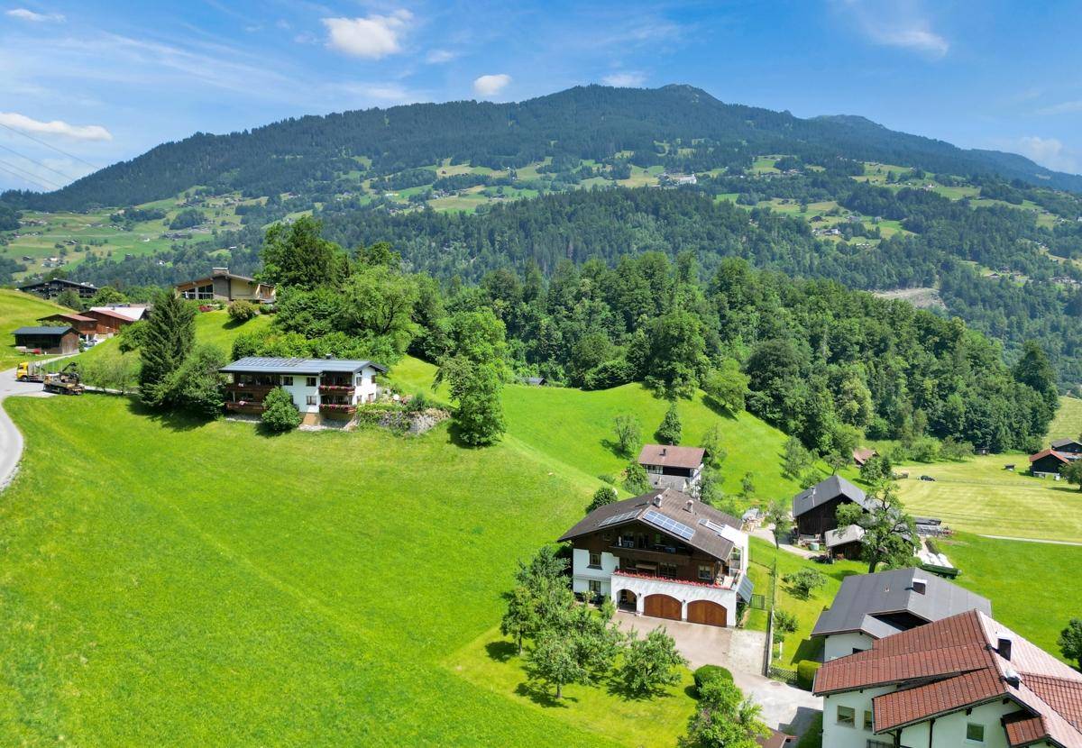 Mehrere Häuser in einer malerischen Berglandschaft mit grünen Hängen und Wäldern.