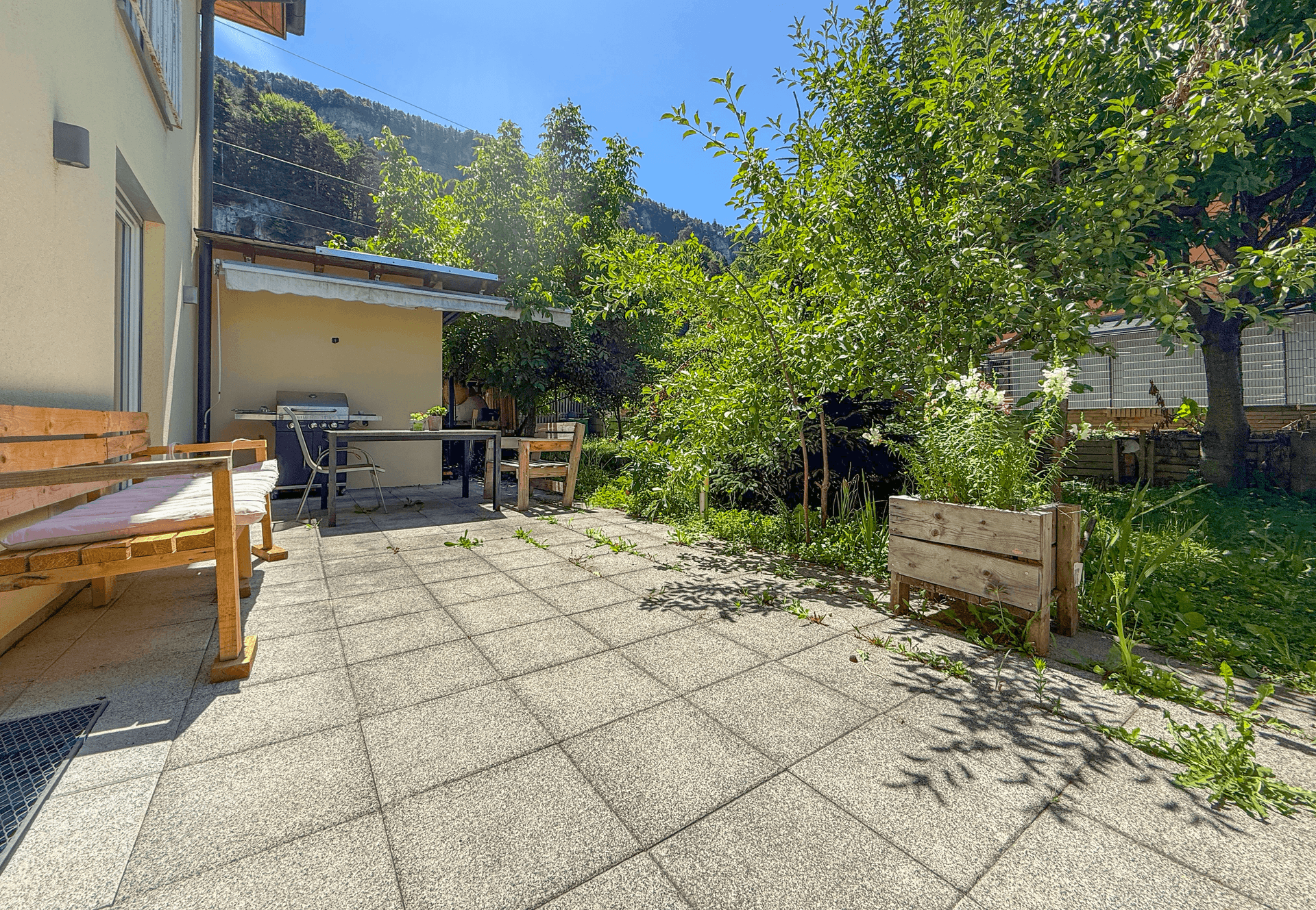 Gepflasterte Terrasse mit Grillbereich und Gartenmöbeln, umgeben von Bäumen, ideal für Sommerabende.