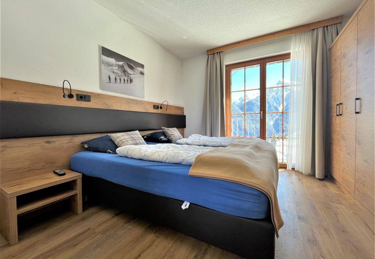 Schlafzimmer mit Holzbett, Kleiderschrank und Balkonzugang mit Bergblick.