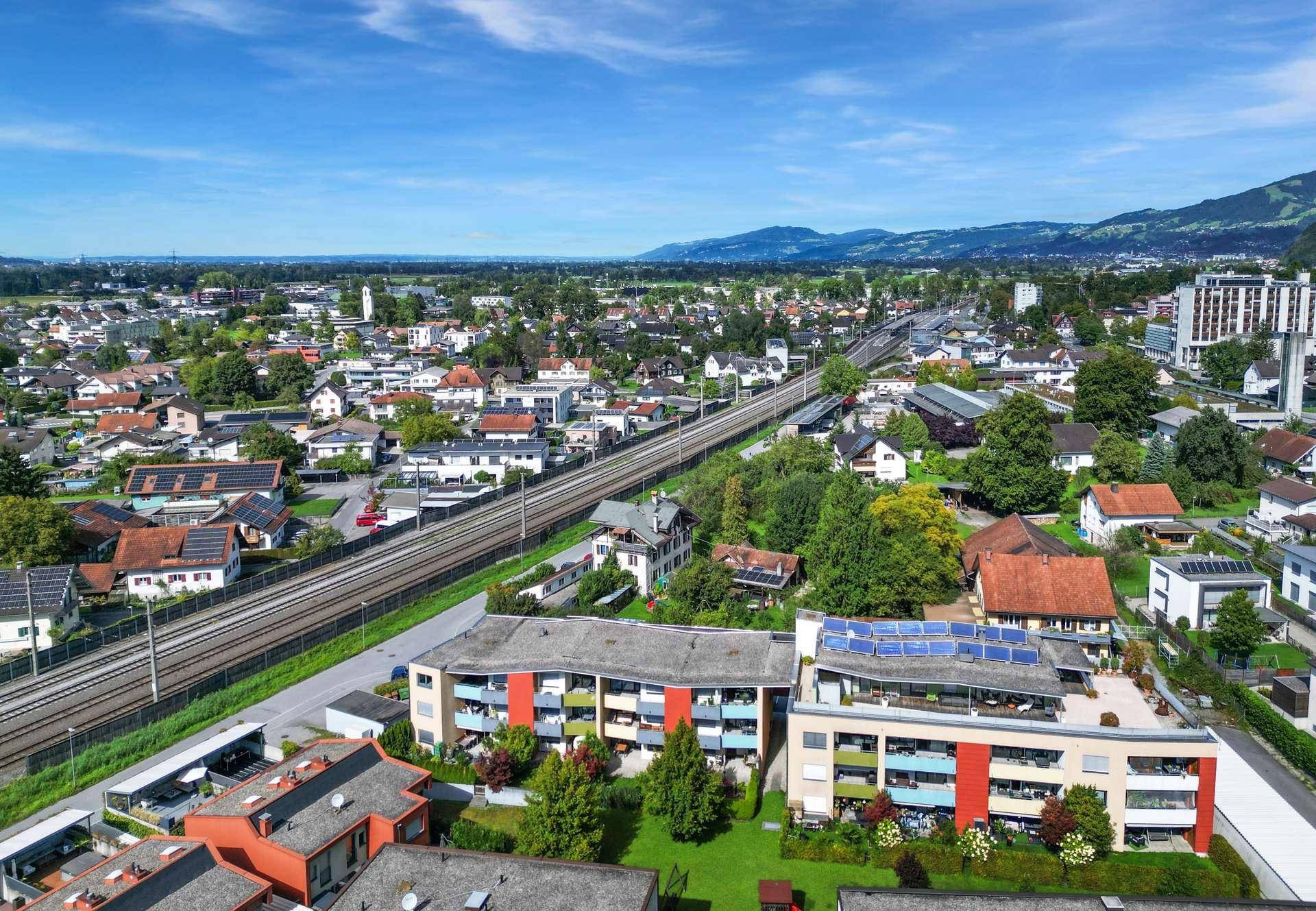 Weitläufige Luftaufnahme der Wohngegend mit Gebäuden, Bahnlinie und Bergen im Hintergrund.