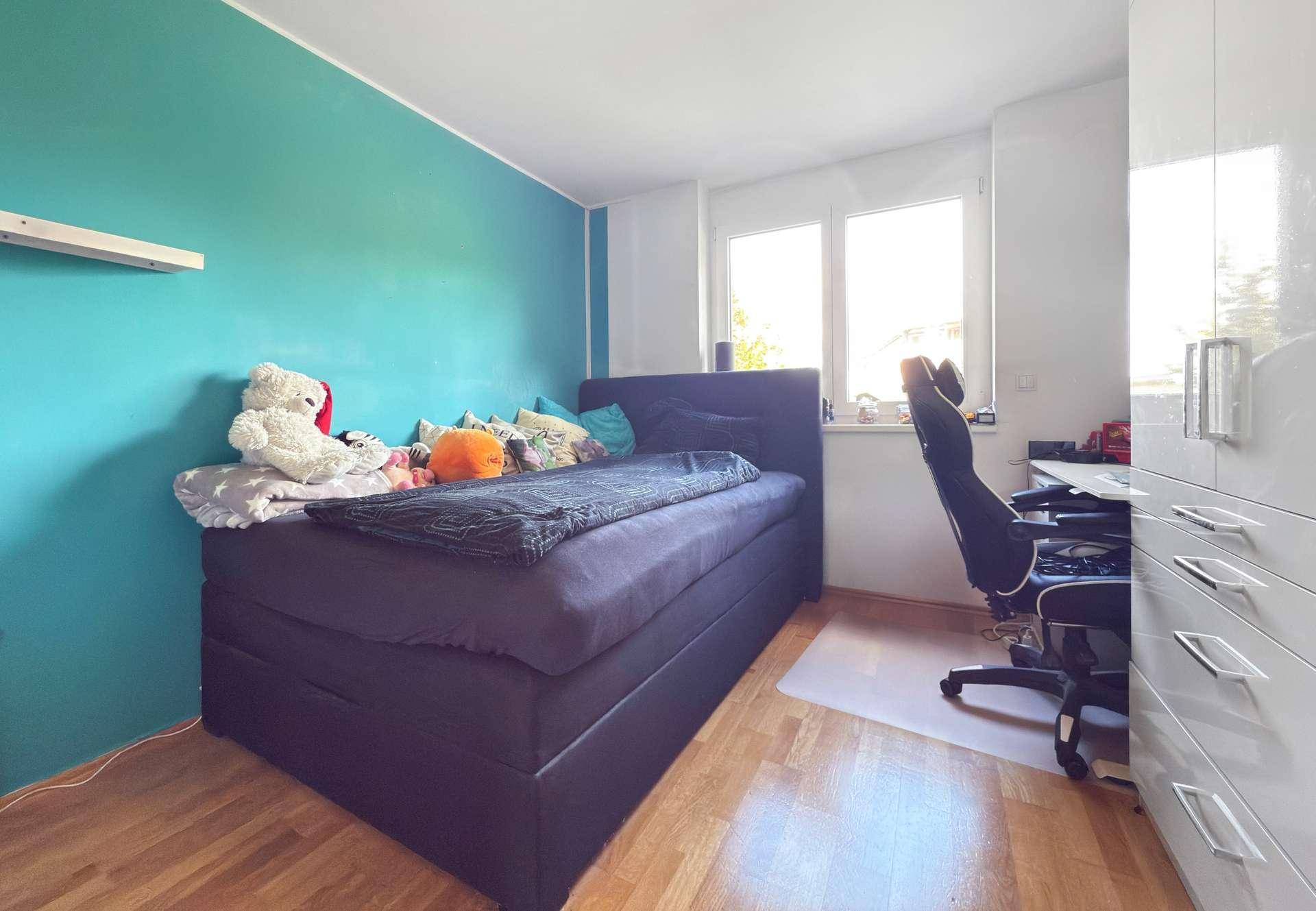 Kinderzimmer mit blaugrüner Wand, Einzelbett und Schreibtisch am Fenster.