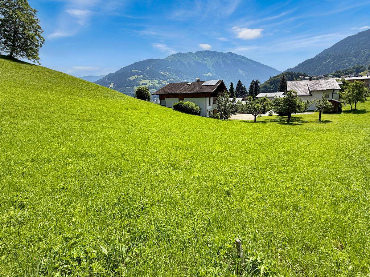 Weitläufige grüne Wiesen mit Häusern und majestätischen Bergen im Hintergrund unter blauem Himmel.