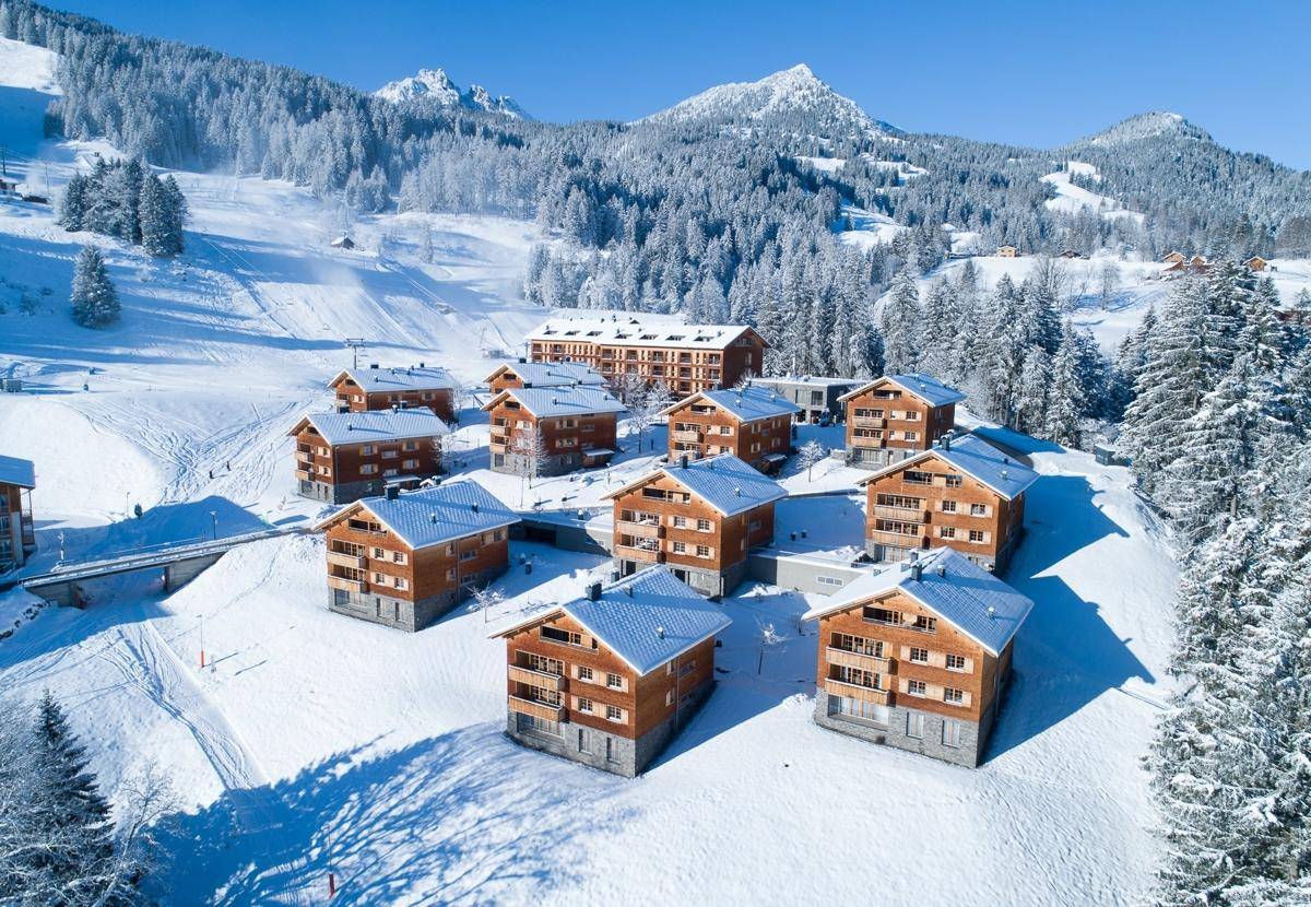 Winterliche Luftaufnahme einer Ferienanlage mit Chalets und schneebedeckten Bergen.